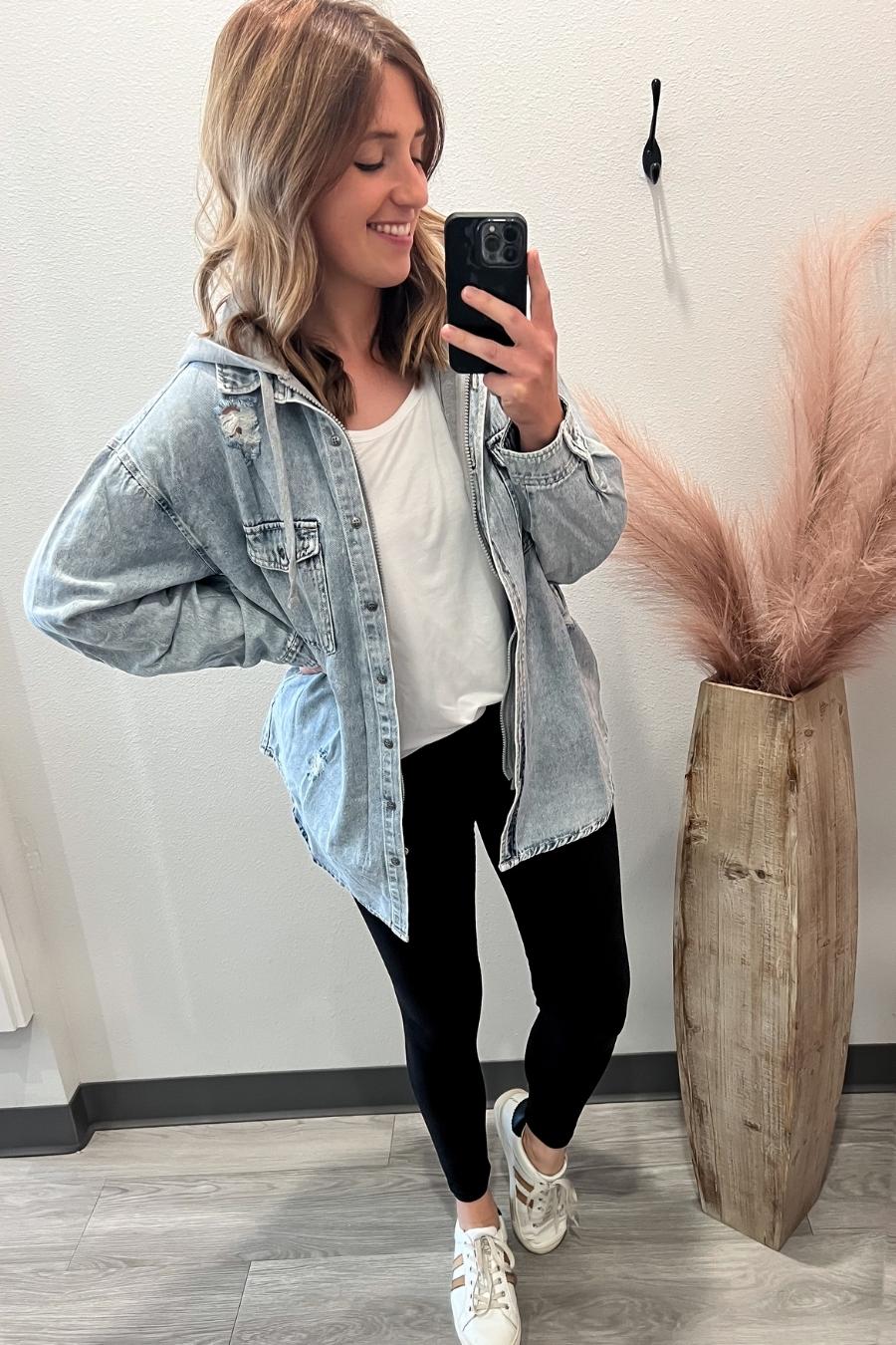 Woman taking a selfie wearing a denim jacket, white shirt, and black pants in a room with a vase of pink grasses.