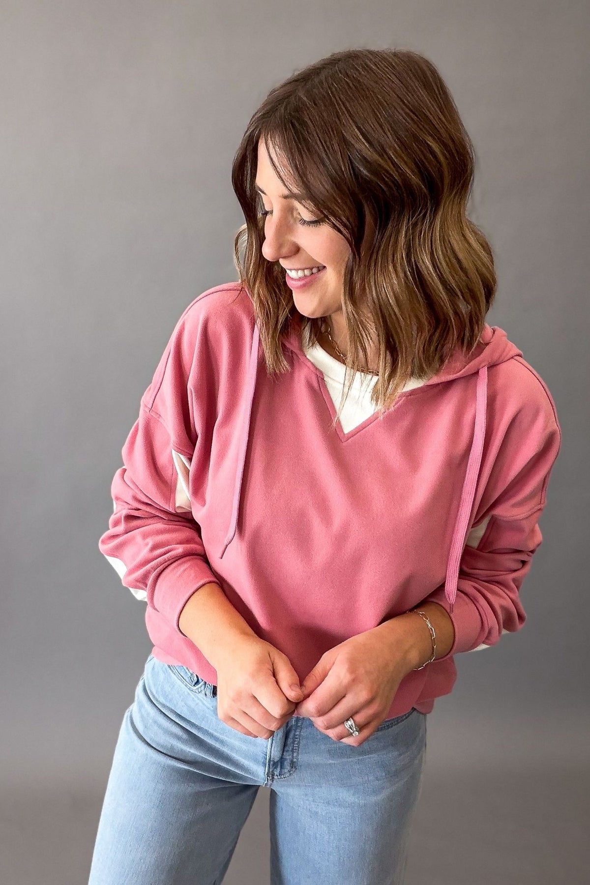 A woman wearing a dusty pink pullover hoodie with white side panels and light blue jeans, standing against a solid gray studio background.