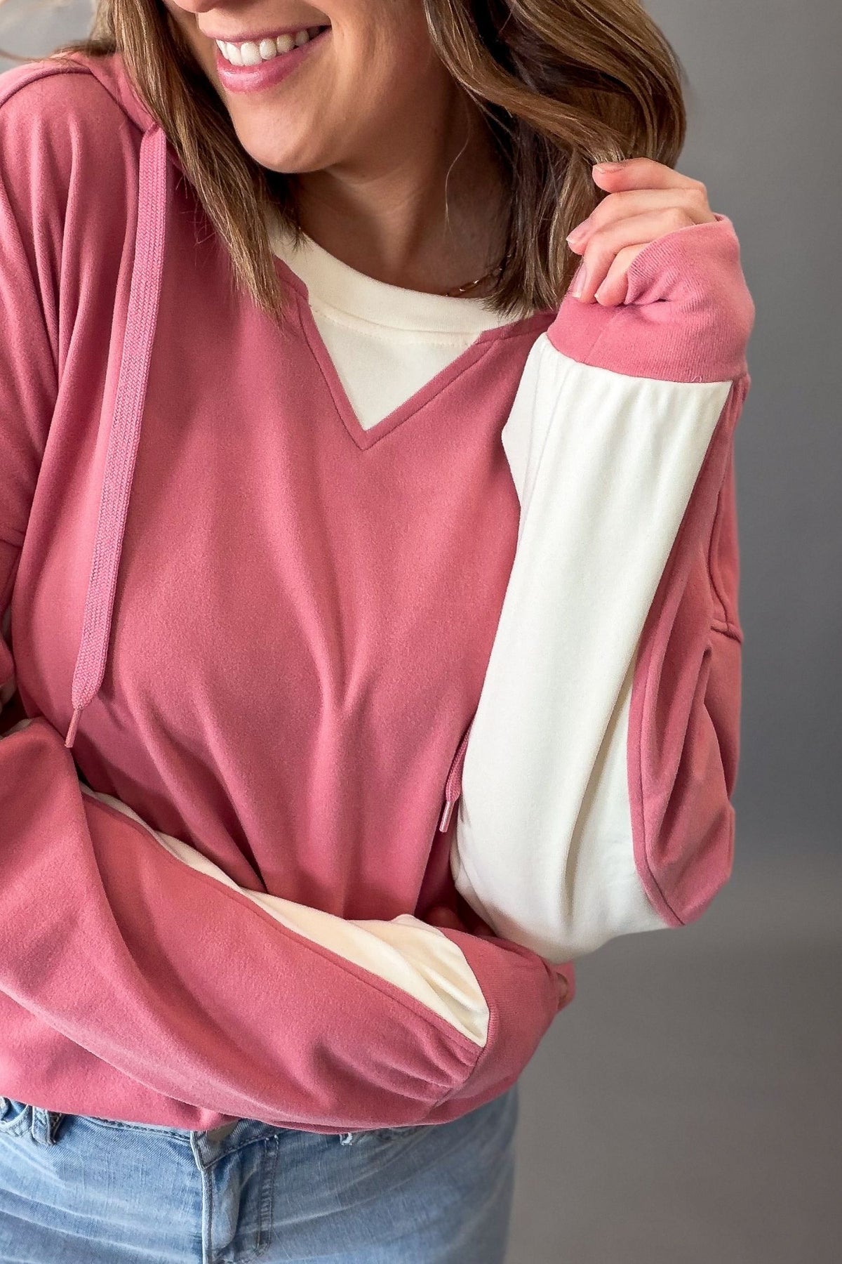 A woman wearing a dusty rose pink color-block hoodie with cream-colored inner sleeves and a cream V-neck undershirt, paired with blue jeans.