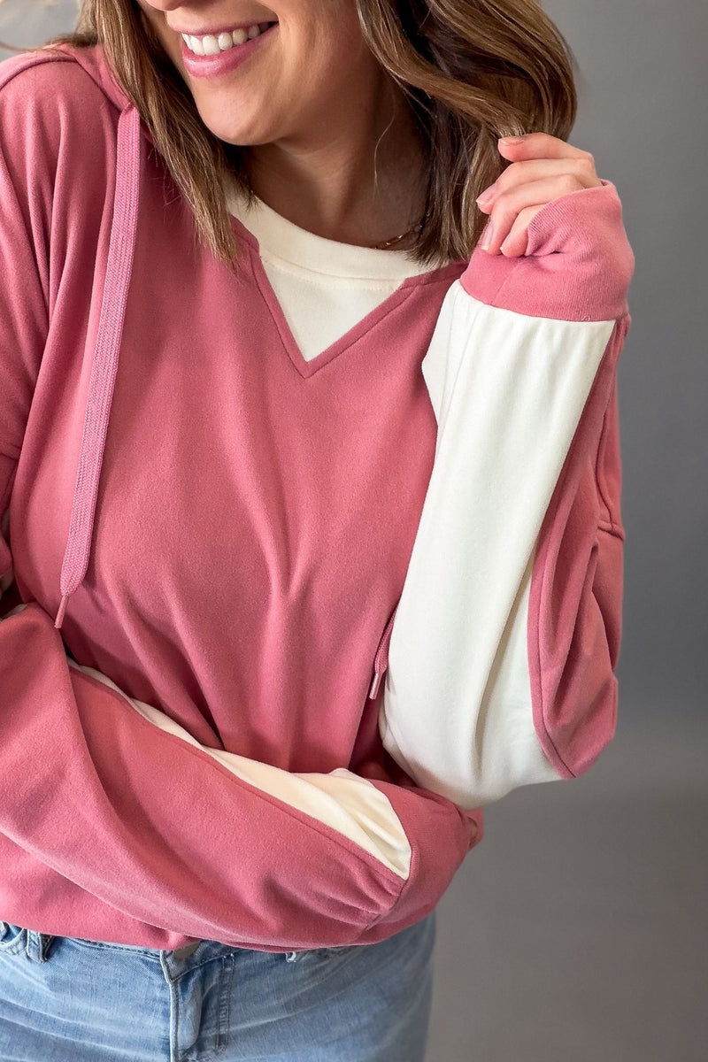 A woman wearing a dusty rose pink color-block hoodie with cream-colored inner sleeves and a cream V-neck undershirt, paired with blue jeans.