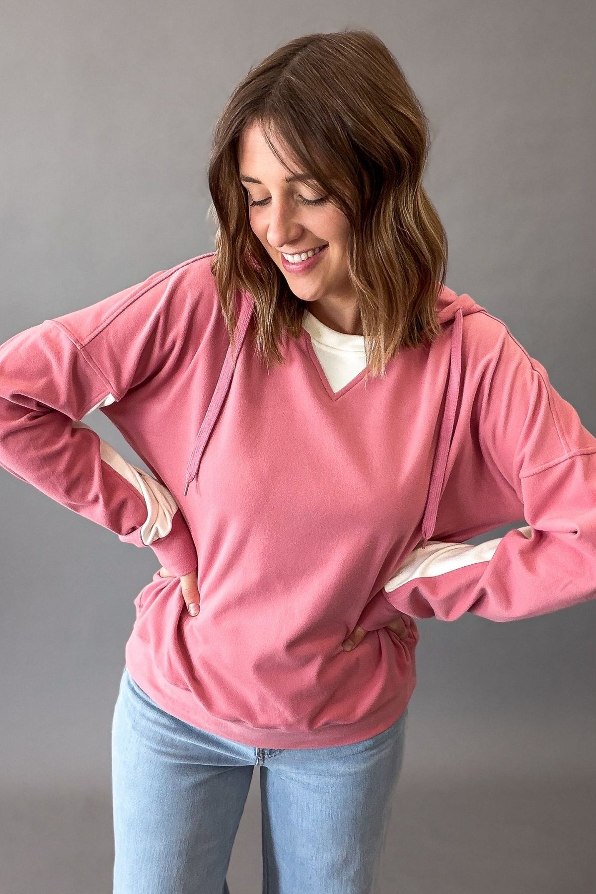 A woman wearing a dusty pink pullover hoodie with white contrast stitching and drawstrings, layered over a white t-shirt and paired with light blue jeans, posing against a neutral gray background.