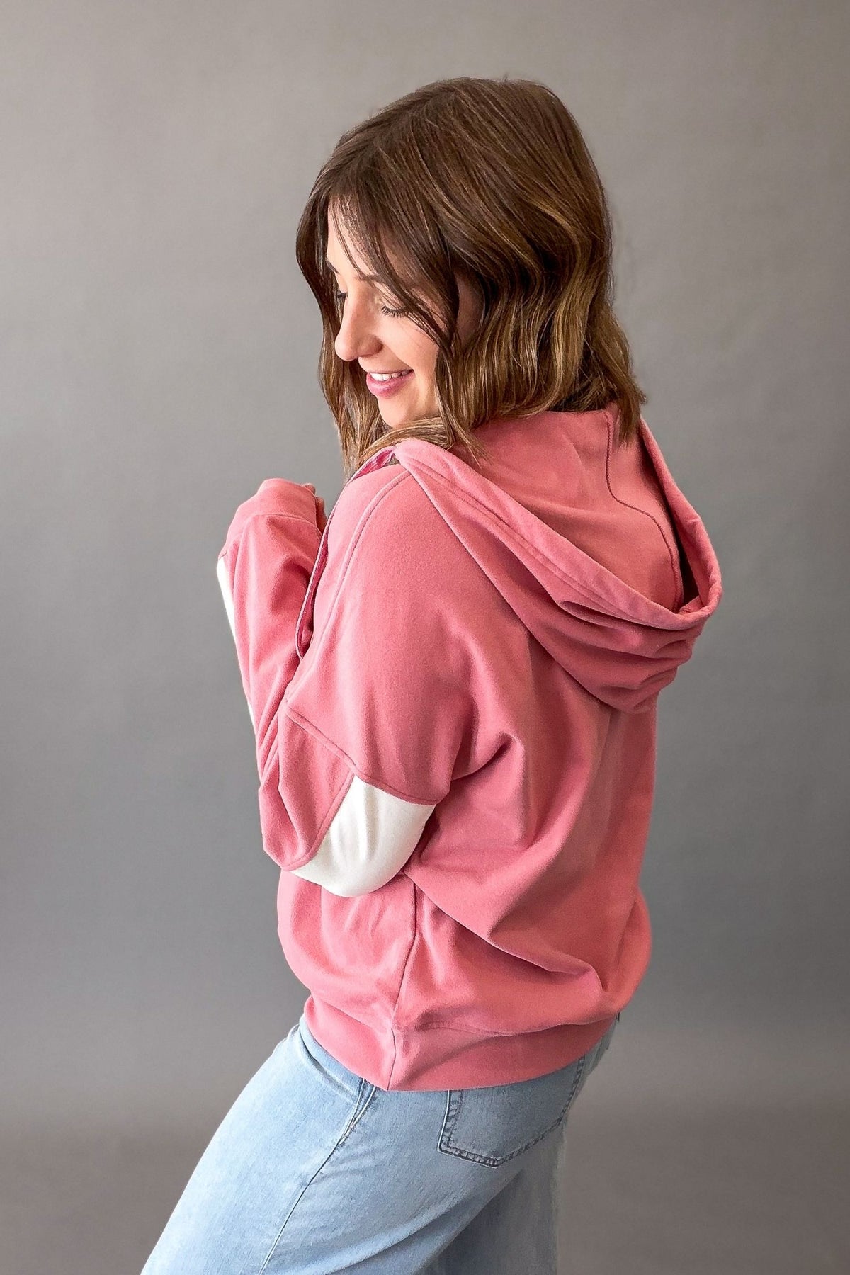 A woman wearing a dusty pink hoodie with contrasting white elbow patches, paired with light blue jeans, photographed against a neutral gray studio background.
