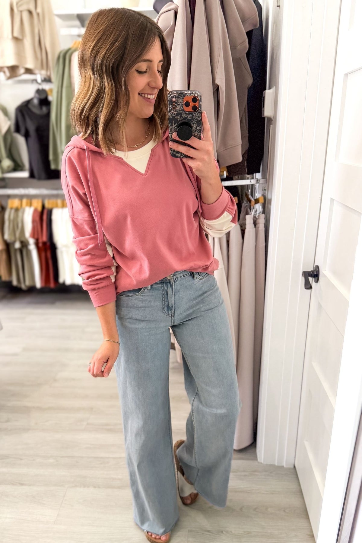 A woman takes a mirror selfie in a clothing boutique, wearing a dusty pink long-sleeve hoodie over a cream V-neck top and light-wash wide-leg jeans.
