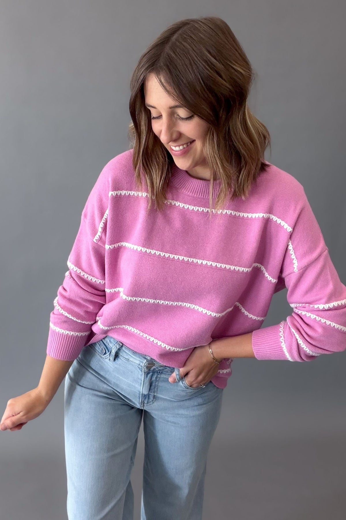 A woman wearing a pink knit sweater with white horizontal wavy knit stripes and light blue jeans, standing against a solid gray studio background.