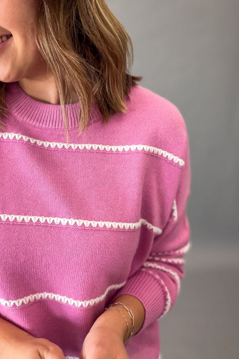 A close-up of a woman wearing a pink knit sweater with white horizontal striped patterns, featuring her hair, neck, and hands visible against a neutral gray background.