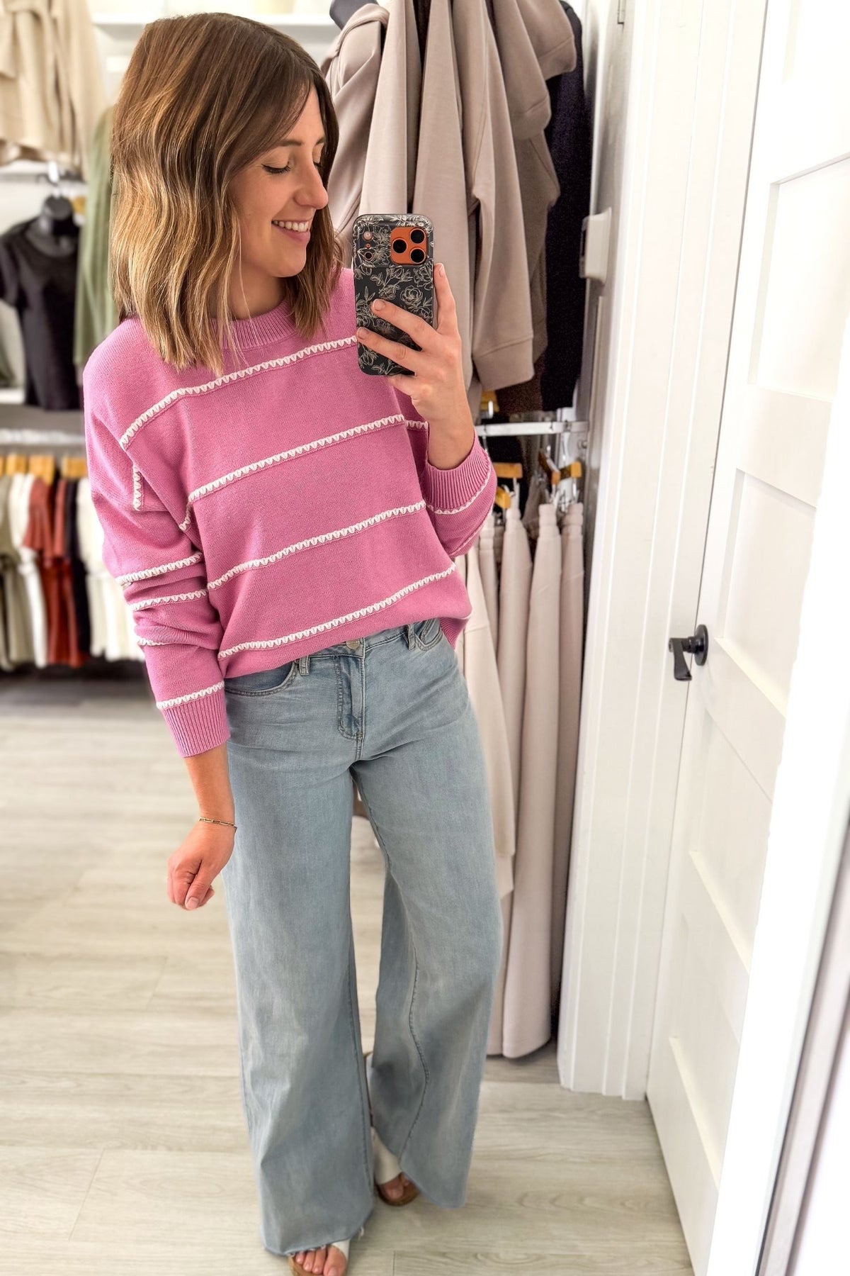 A woman takes a mirror selfie in a clothing boutique, wearing a pink knit sweater with white horizontal stripes and light-wash wide-leg jeans.