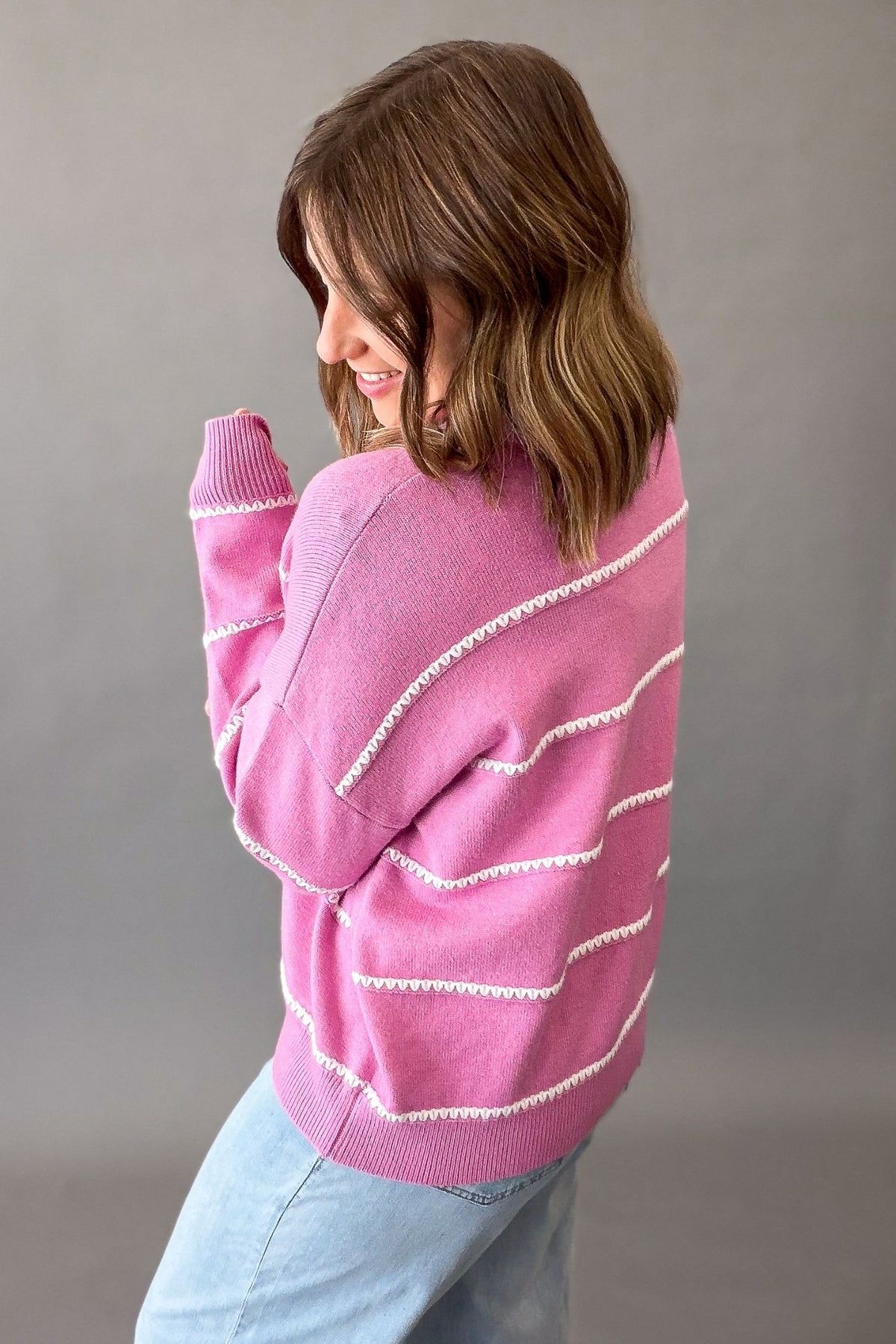 A woman wearing a pink knit sweater with white textured horizontal stripes, paired with light blue jeans, photographed against a neutral gray studio background.