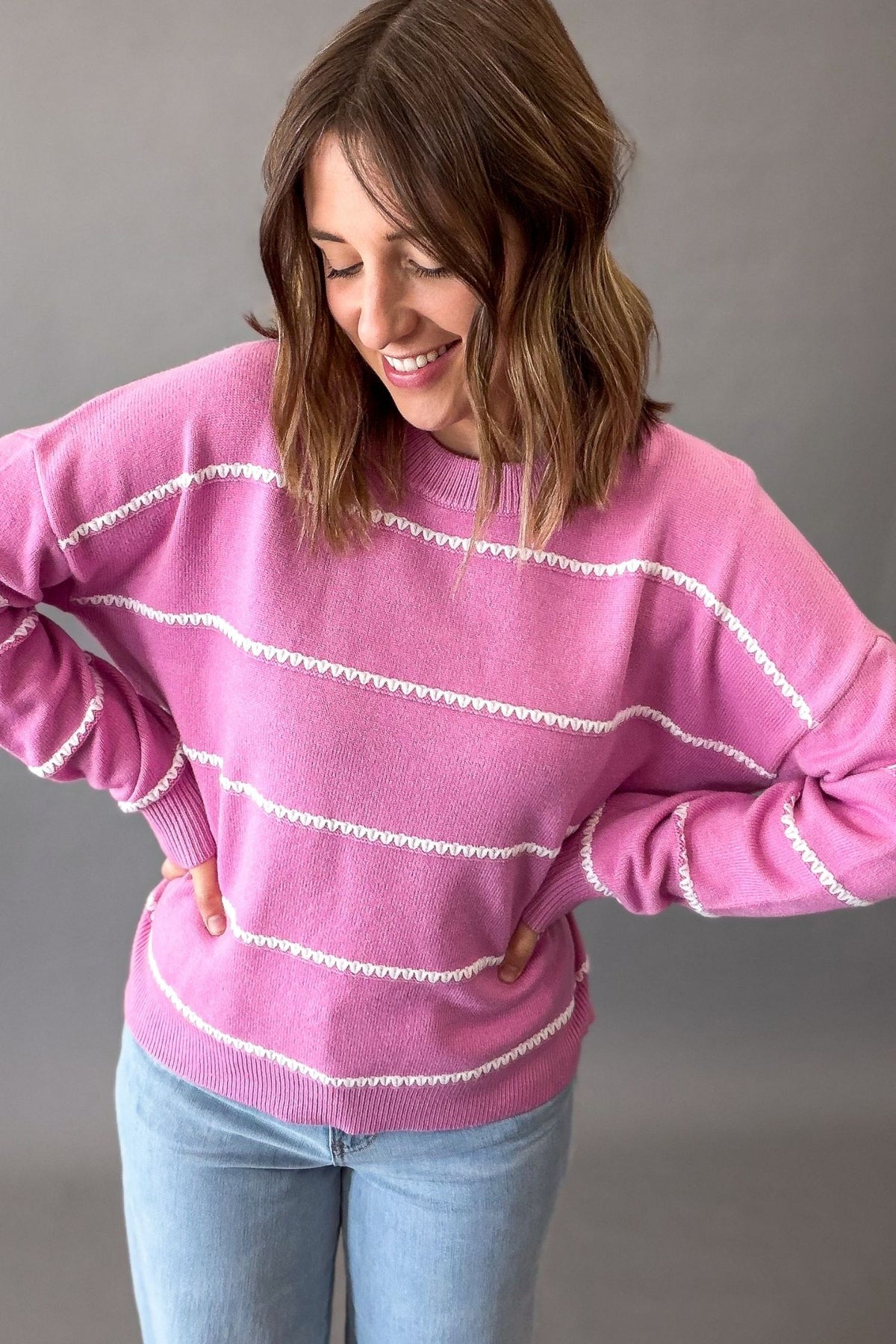 A woman wearing a pink knit sweater with white textured horizontal stripes and light blue jeans, standing against a plain gray studio background.