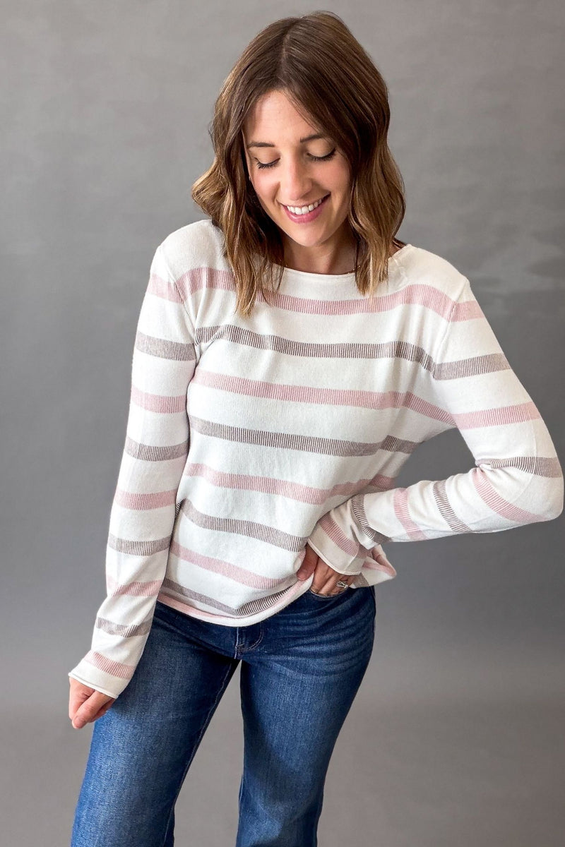 A woman wearing a long-sleeve knit sweater with horizontal stripes in white, pink, and mocha, paired with blue jeans, standing against a neutral gray studio background.
