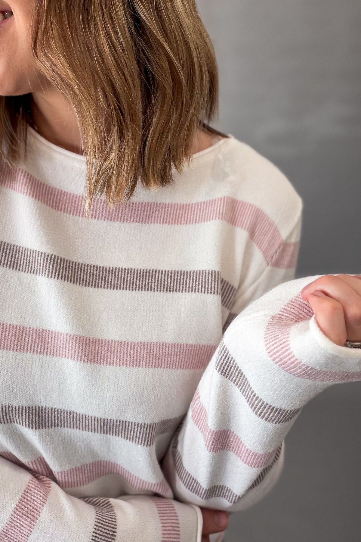 A close-up of a person wearing a soft knit sweater with horizontal stripes in pink and mocha on an off-white base, against a plain gray background.