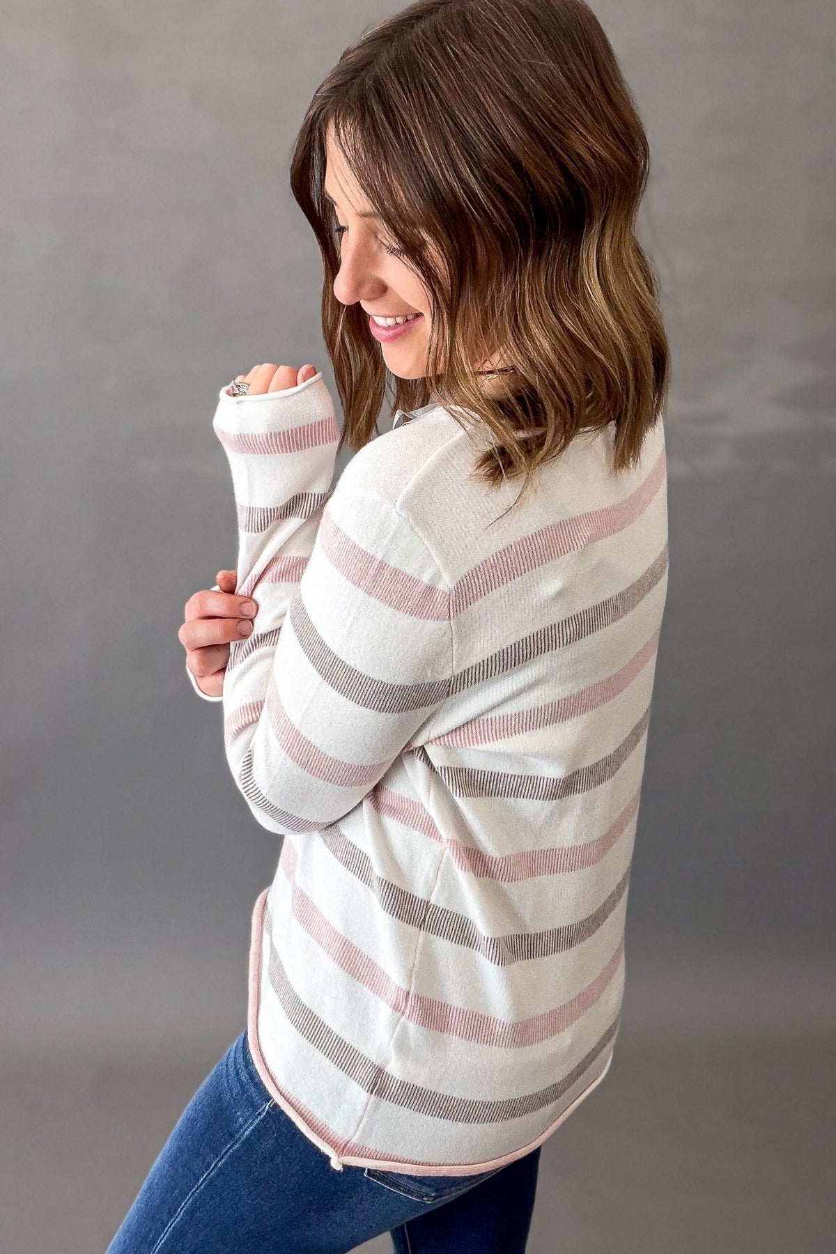 A woman wearing a cream-colored knit sweater with horizontal pink and mocha stripes, paired with blue jeans, posing against a neutral gray studio background.
