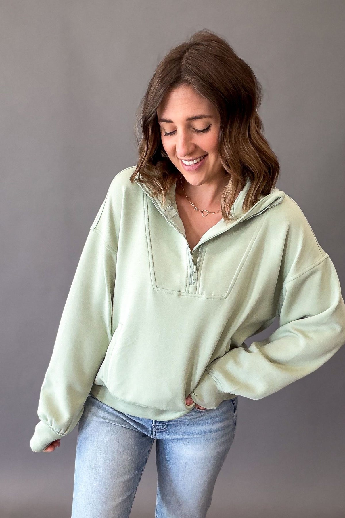woman wearing a light green, pistachio, quarter zip and blue jeans on a grey background