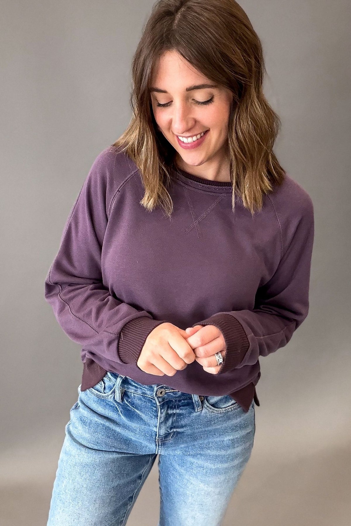 A woman wearing a dusty purple long-sleeve crewneck sweatshirt and blue jeans, standing against a neutral gray studio background.