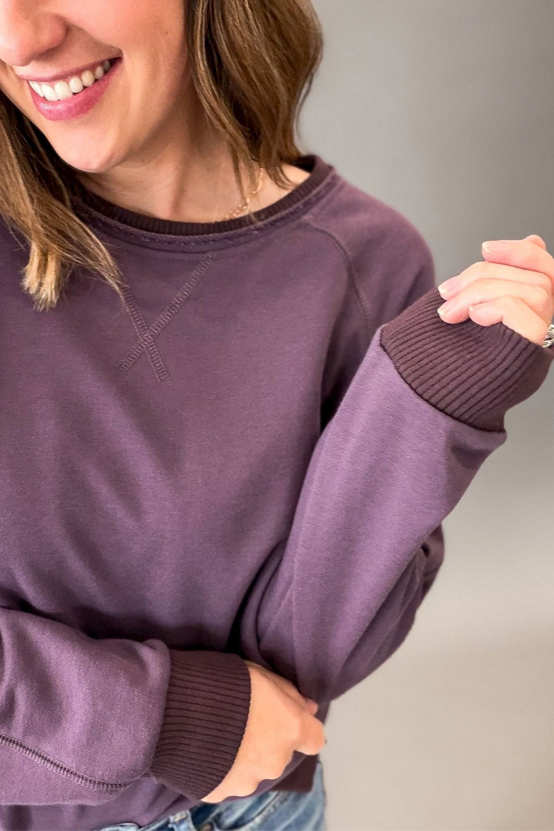 A close-up of a woman wearing a dusty purple crewneck sweatshirt with ribbed cuffs and collar, paired with blue jeans against a neutral gray background.