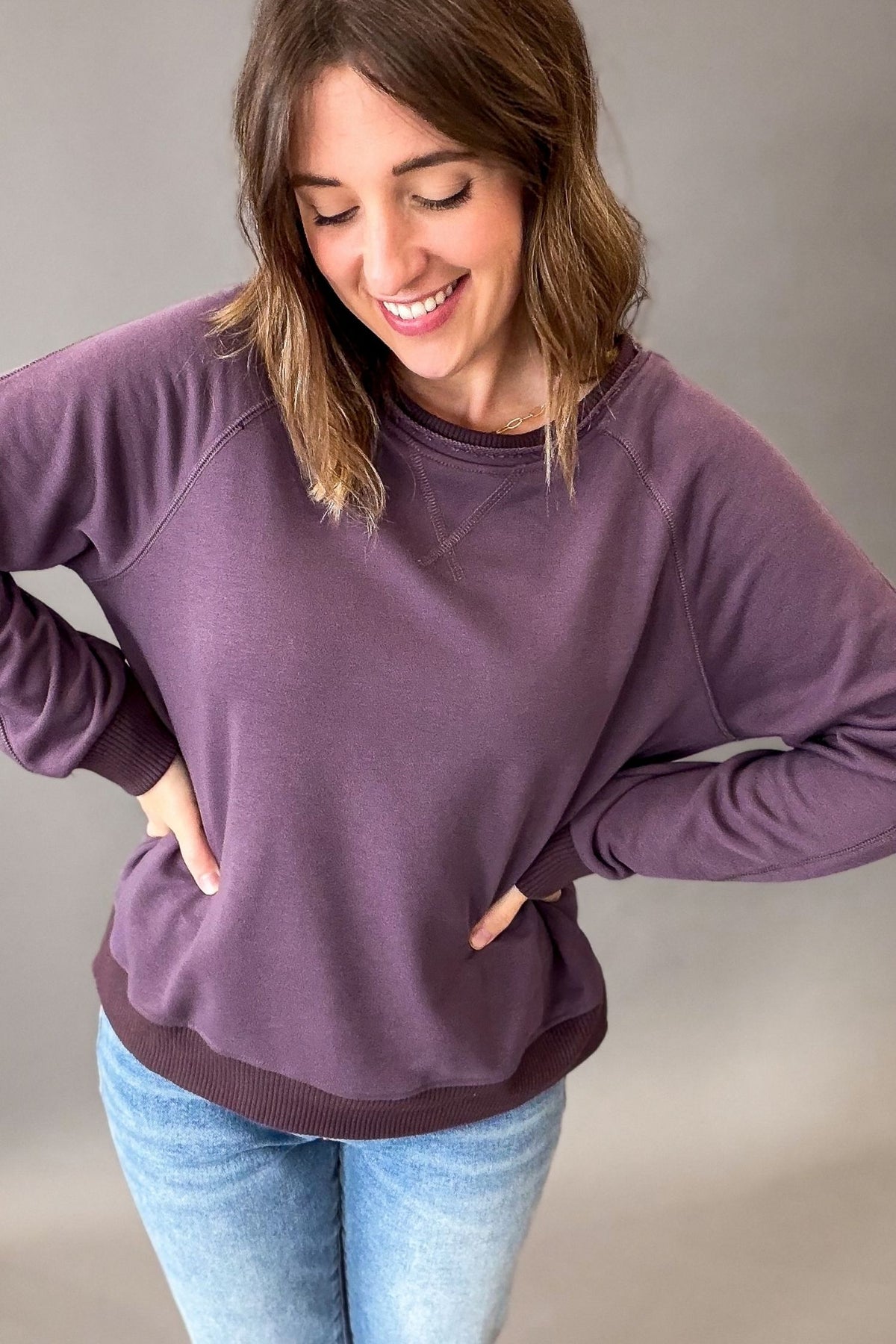 A woman wearing a dusty purple crewneck sweatshirt with side pockets and ribbed cuffs and hem, paired with light blue jeans, posing against a neutral gray studio background.