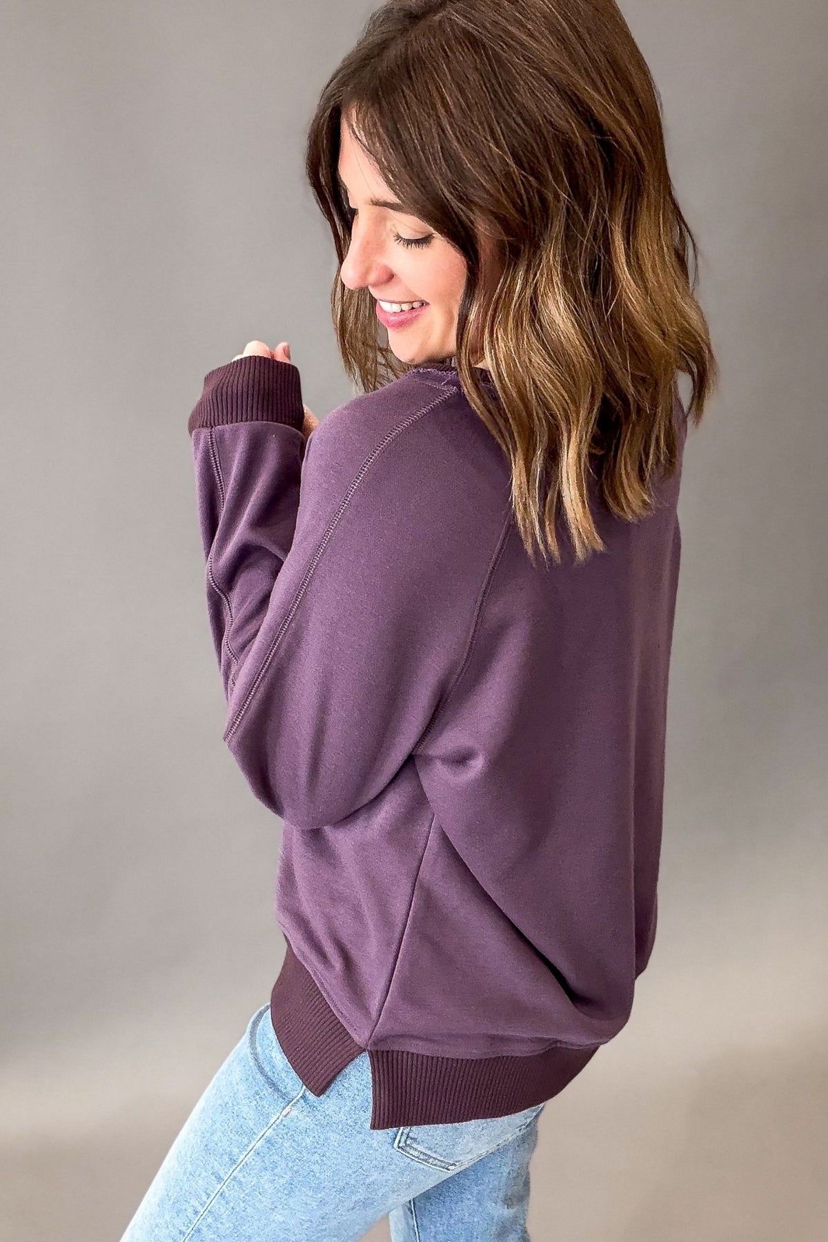 A woman wearing a purple long-sleeve crewneck sweatshirt and light blue jeans, shown from a side-back angle against a neutral gray studio background.