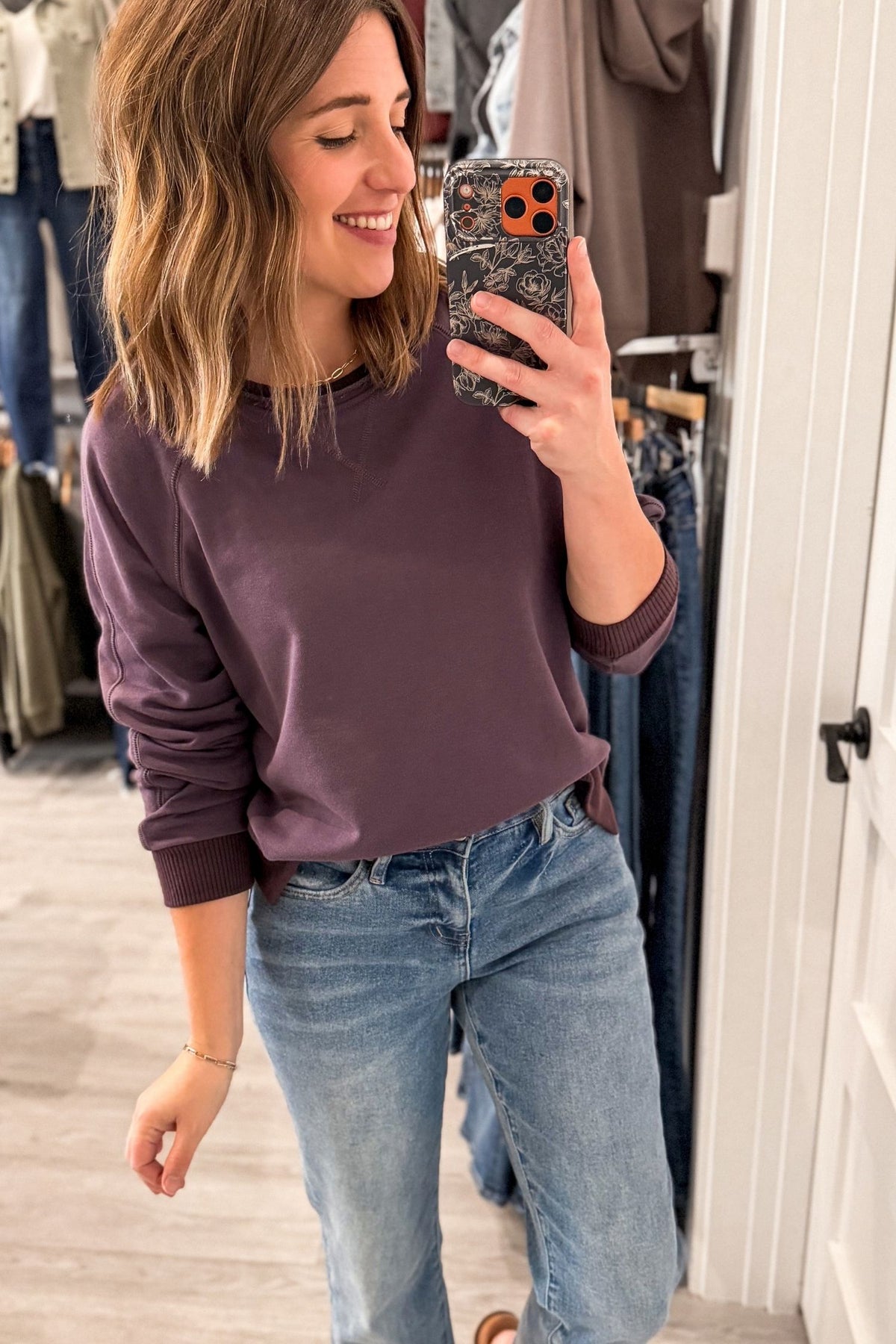 A woman takes a mirror selfie in a clothing store, wearing a dark plum purple long-sleeve crewneck sweatshirt and light wash blue jeans.