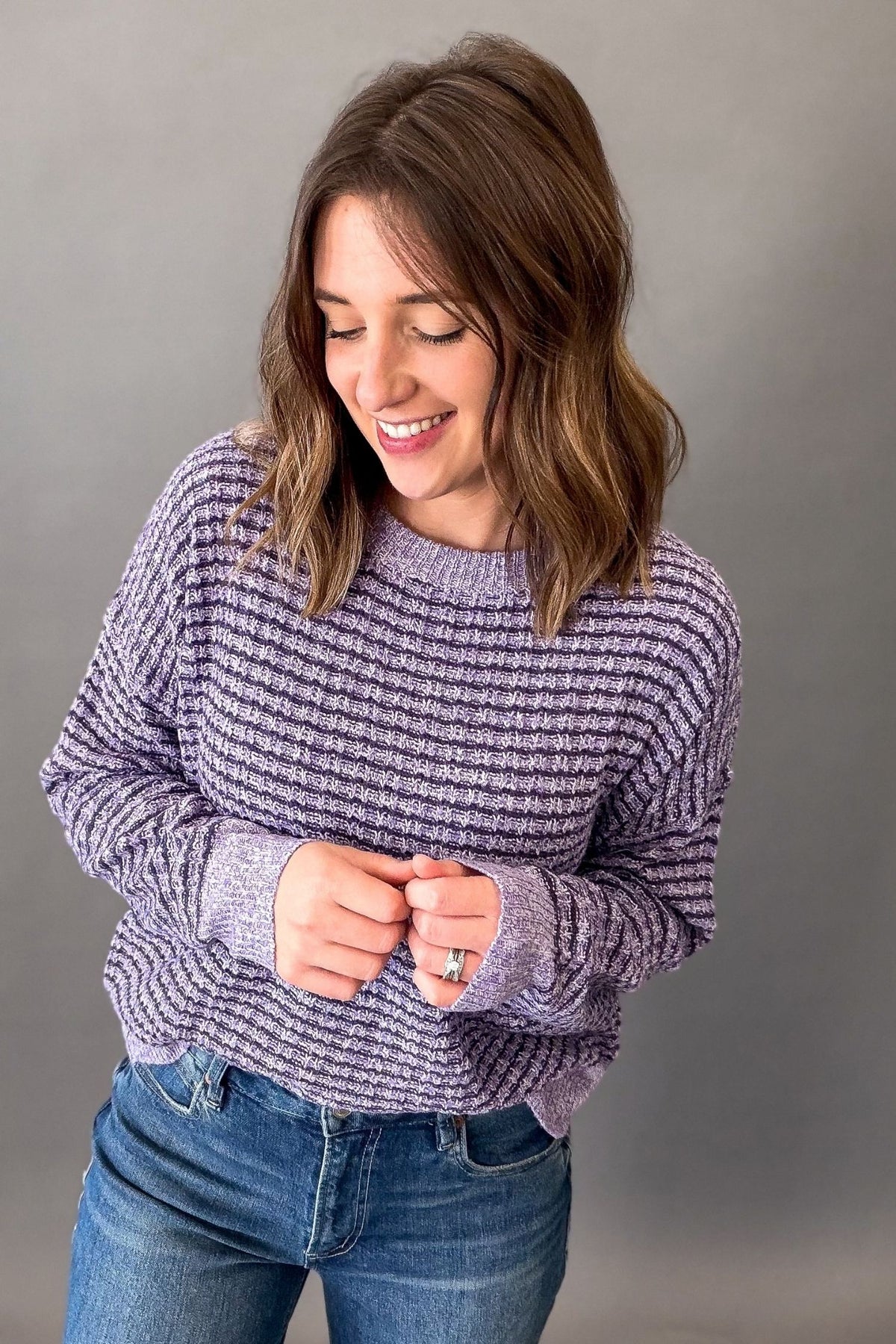 A woman with shoulder-length brown hair smiling and looking down, wearing a textured lavender and purple knit sweater with blue jeans, standing against a solid gray studio background.