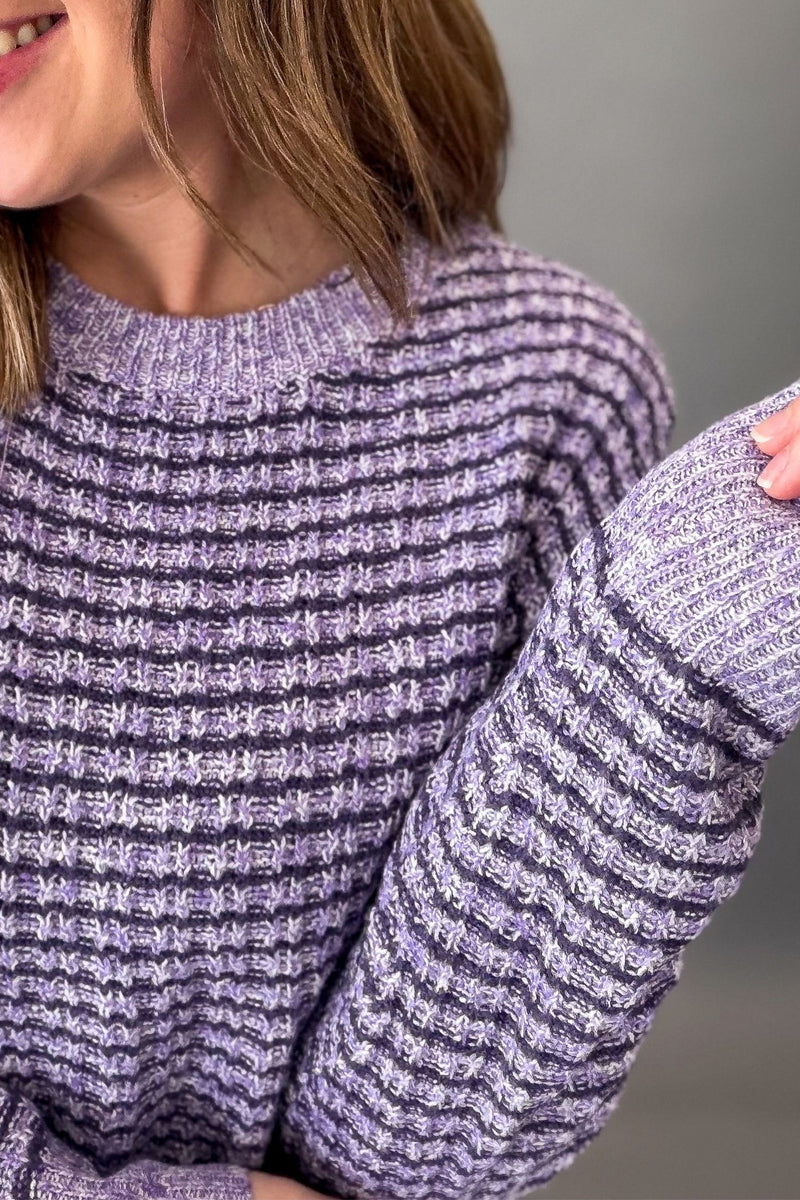 A close-up of a person wearing a chunky knit crewneck sweater in a heathered lavender and dark gray colorway, showing the ribbed texture and sleeve details.