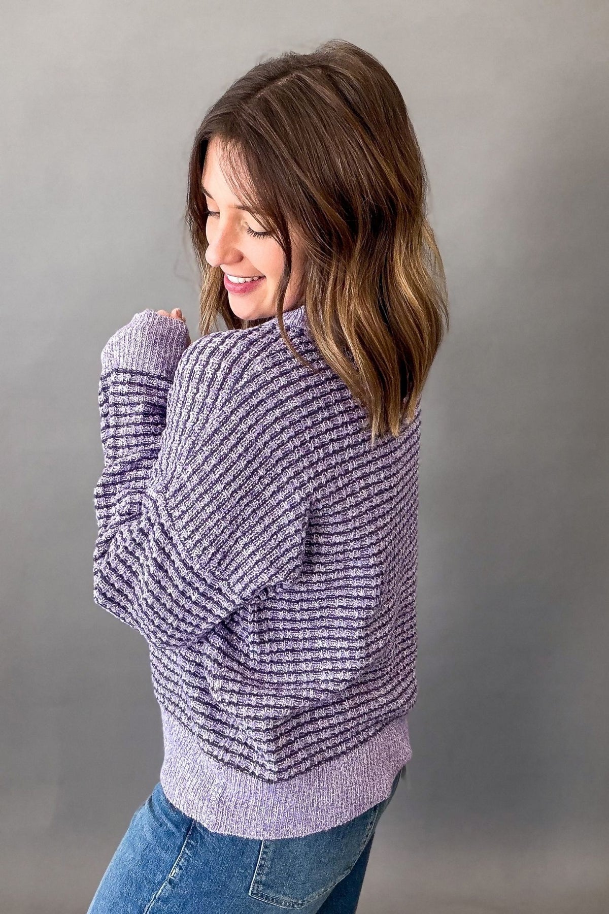 A woman wearing a lavender and purple marled ribbed knit sweater, shown from a side-back angle against a neutral gray studio background.