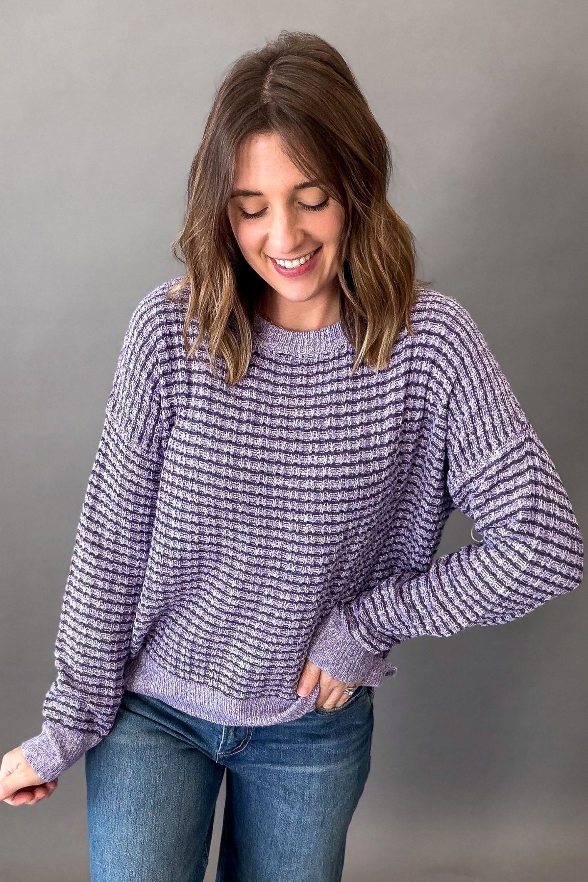 A woman wearing a lavender and purple textured knit sweater with blue jeans, standing against a solid gray studio background.