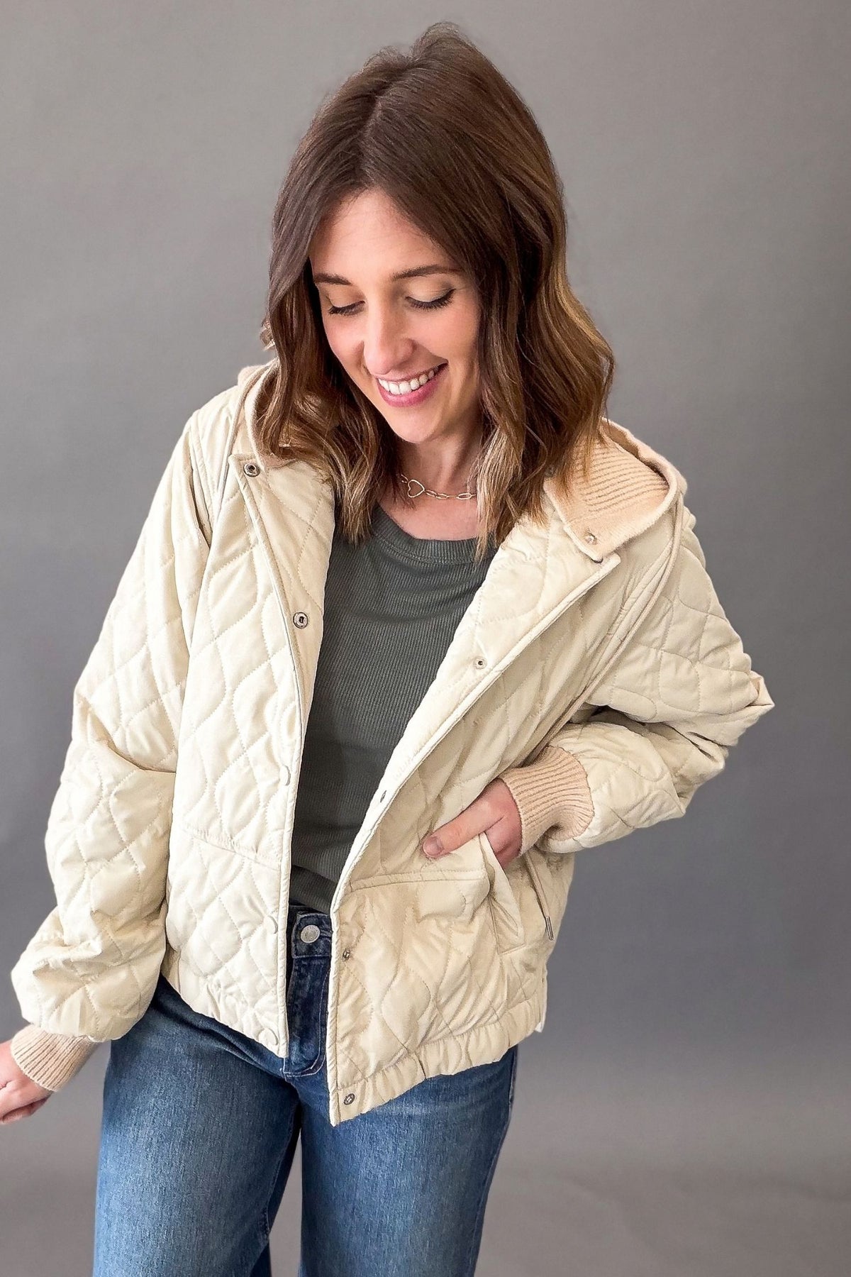 A woman wearing a cream-colored quilted jacket with a ribbed collar, cuffs, and hem, styled over a dark gray t-shirt and blue jeans against a solid gray studio background.
