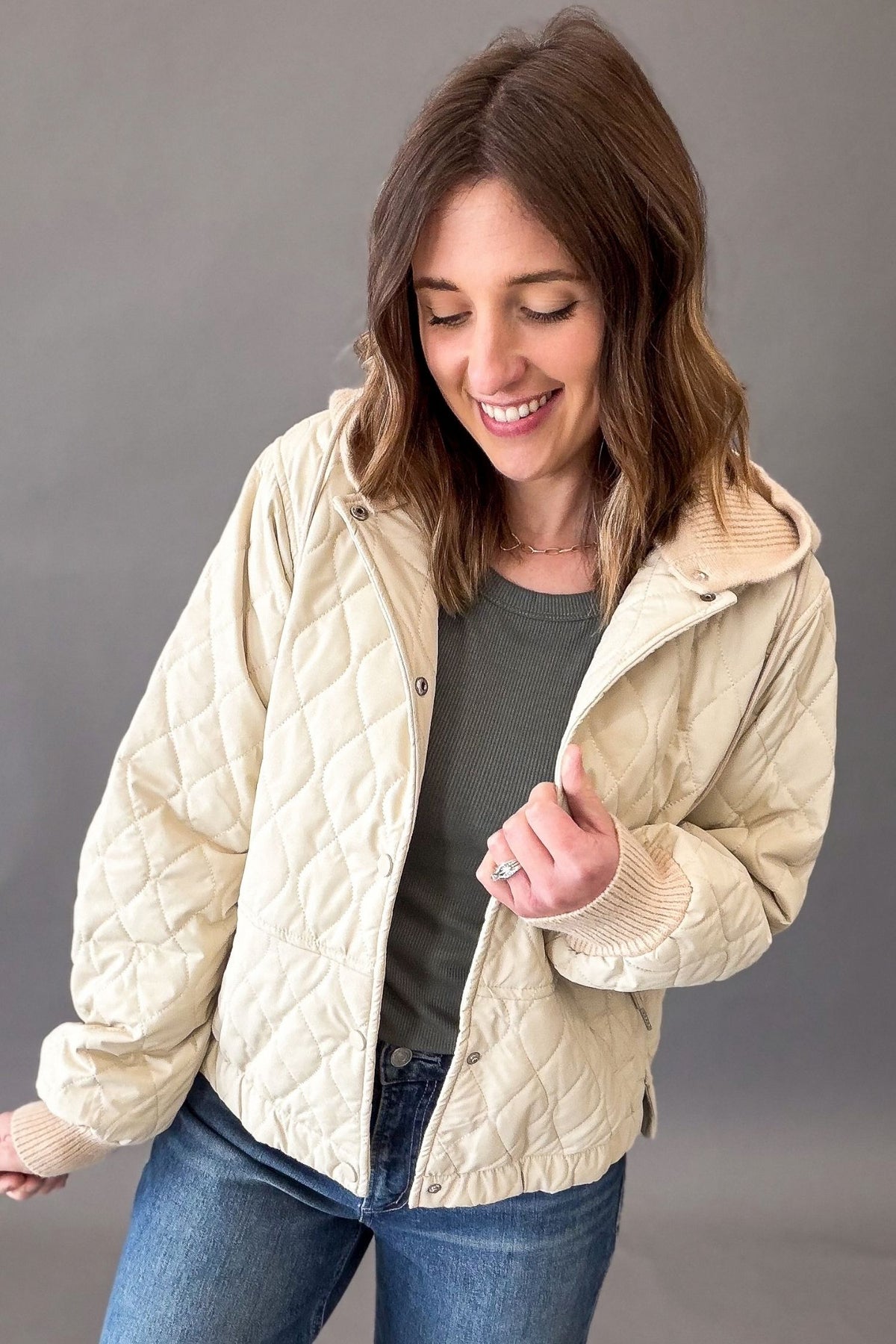 A woman wearing a quilted cream-colored jacket over a dark green long sleeve and blue jeans, posing against a solid gray studio background.