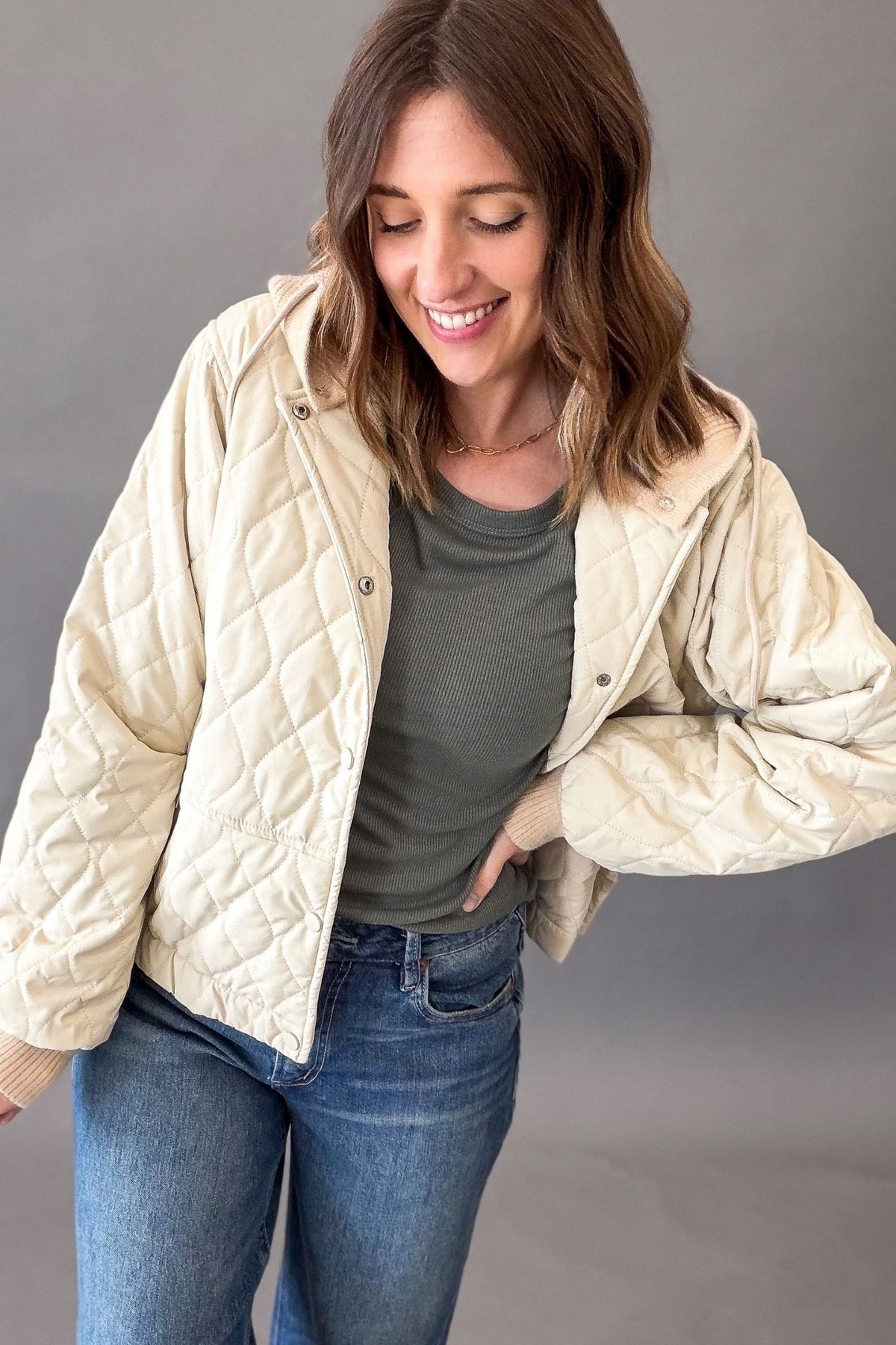 A woman wearing a natural quilted jacket over a dark green long sleeve and blue jeans, posing against a solid gray studio background.