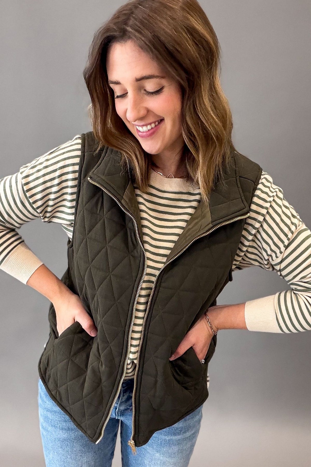 A woman wearing an olive green diamond-quilted zip-front vest over a cream and navy striped long-sleeve shirt, paired with blue jeans, against a solid gray studio background.