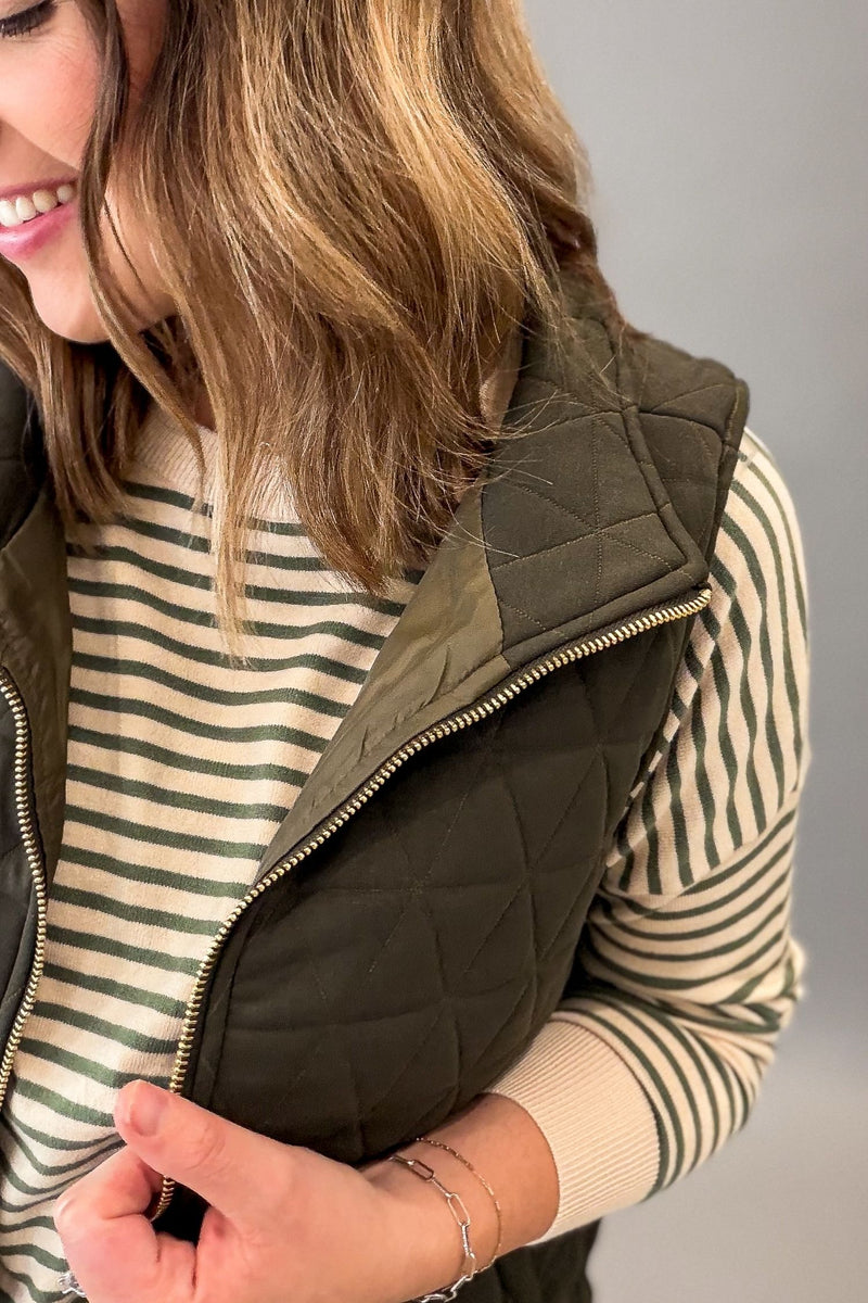 A close-up of a woman wearing an olive green quilted vest over a cream and green striped sweater, holding the vest's zipper.