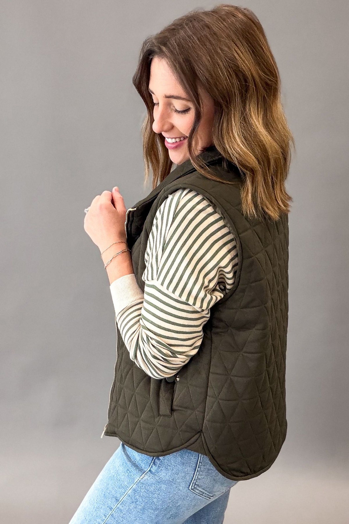 A woman wearing an olive green quilted zip-front vest over a cream and olive striped long-sleeve shirt and light blue jeans, photographed against a plain gray studio background.