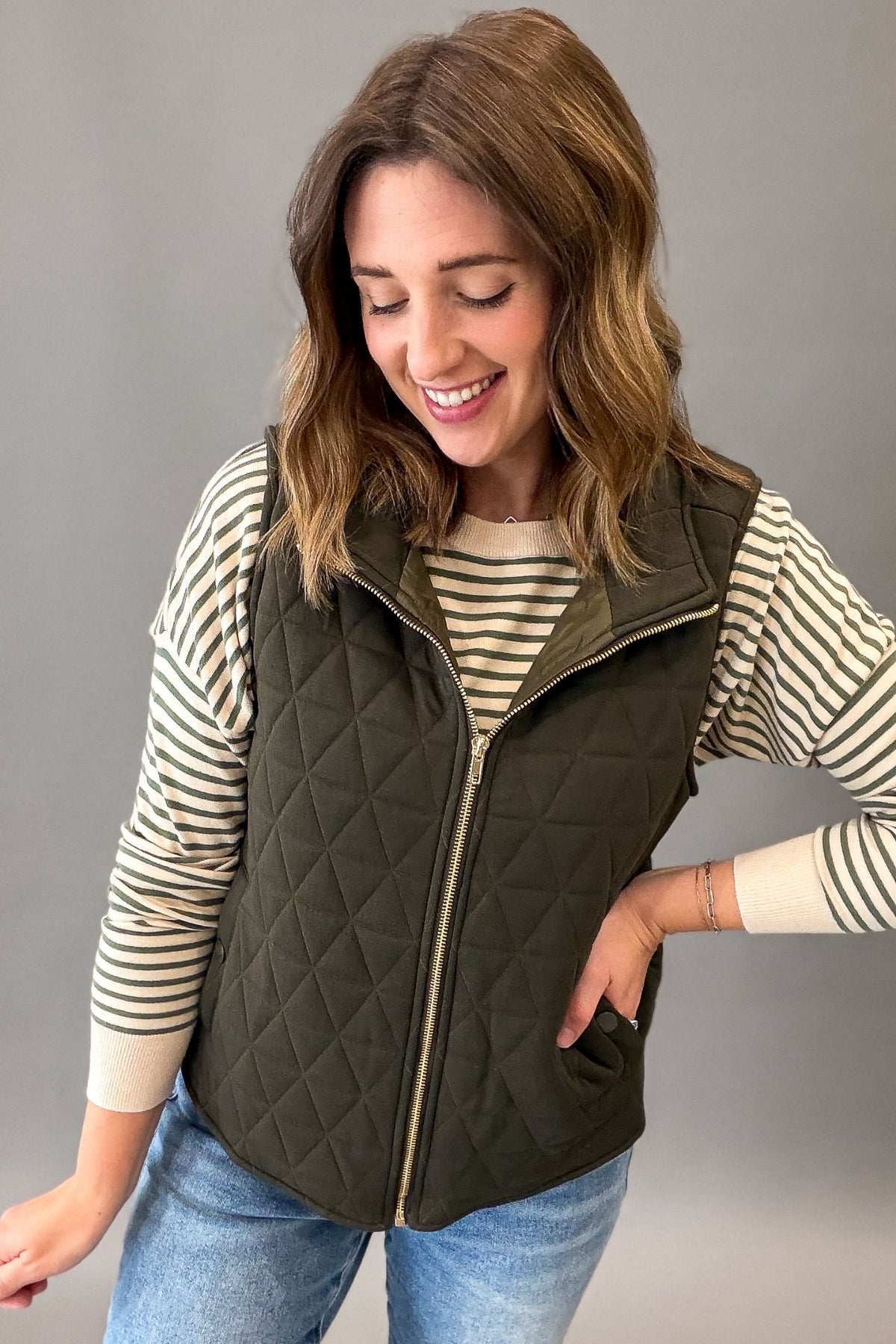 A woman wearing an olive green diamond-quilted zip-up vest over a cream and navy striped long-sleeve shirt, paired with blue jeans, against a solid gray studio background.