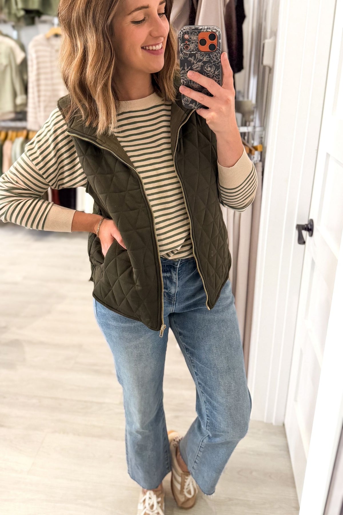 A woman takes a mirror selfie in a boutique while wearing an olive green quilted zip-front vest over a beige and navy striped long-sleeve shirt, paired with light blue jeans and beige sneakers.