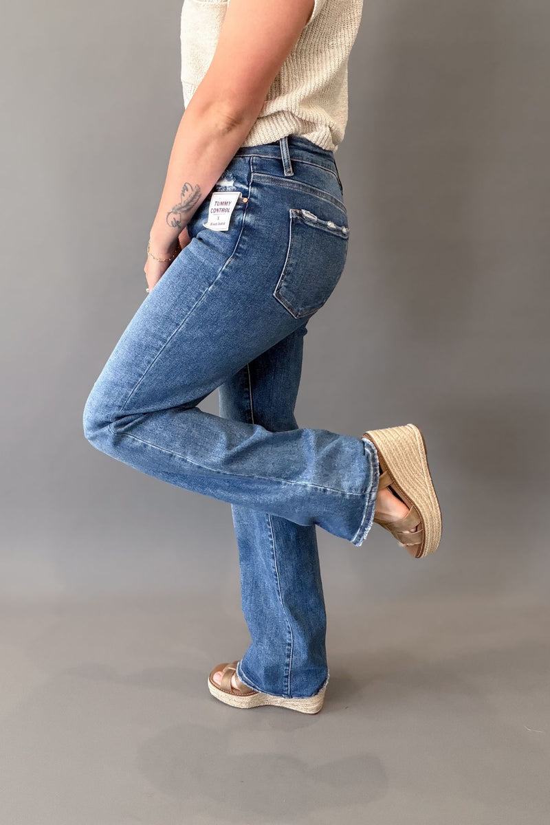A person wearing dark-wash blue straight jeans with a cream ribbed top and neutral wedge sandals, shown from the waist down in a studio setting against a gray background.