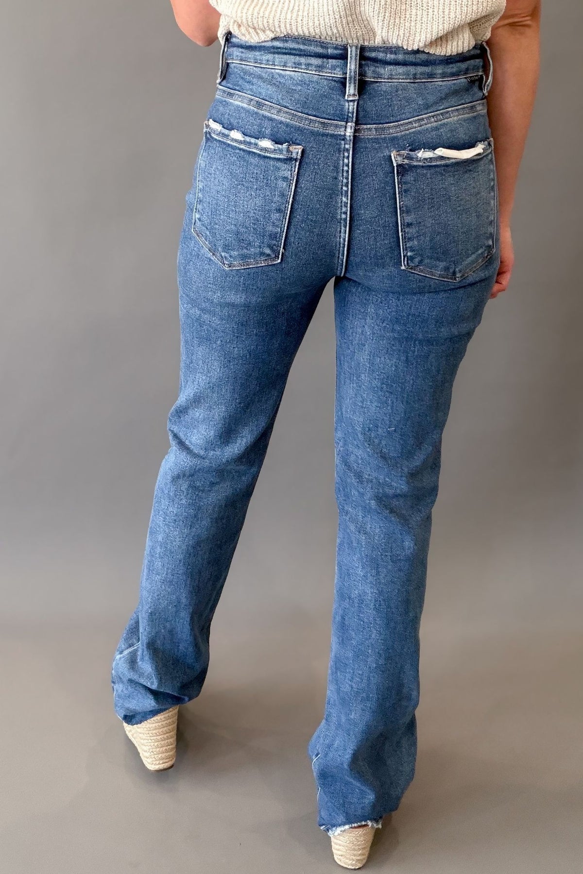 Rear view of a person wearing a dark-wash blue jeans with a straight leg and distressed details on the back pocket flaps, paired with a cream top and beige wedge sandals against a gray studio background.