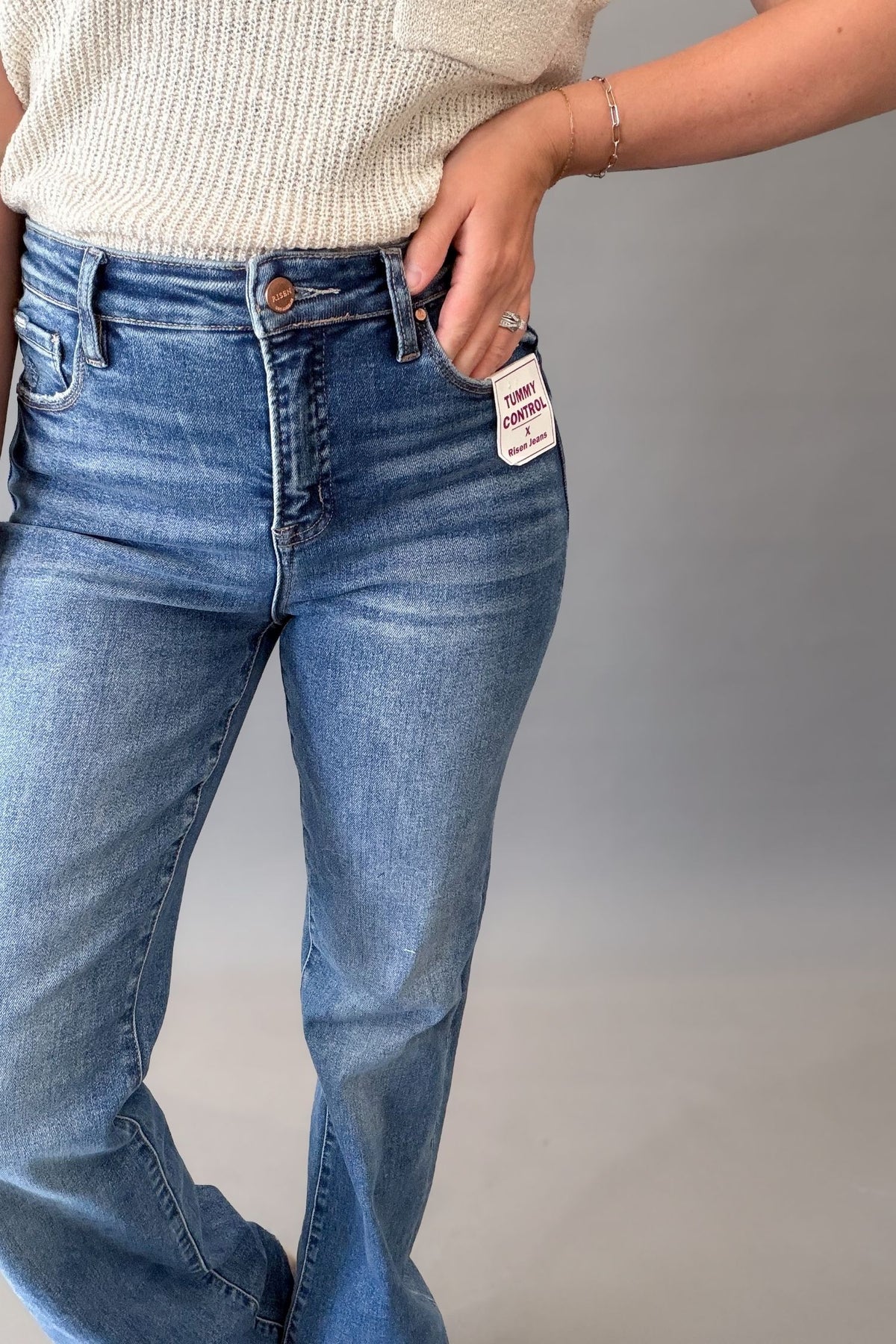 A person wearing dark-wash blue jeans with a cream ribbed knit top, shown from the waist down against a plain gray studio background. A white brand tag is visible on the side pocket.