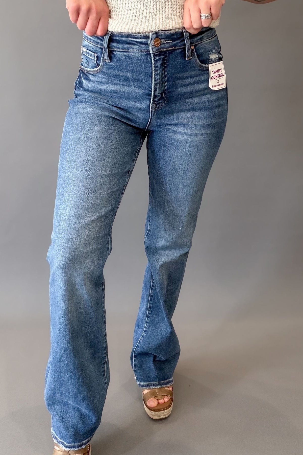 A person wearing dark-wash blue denim jeans with a straight silhouette, paired with a cream knit top and platform sandals, standing against a neutral gray studio background.