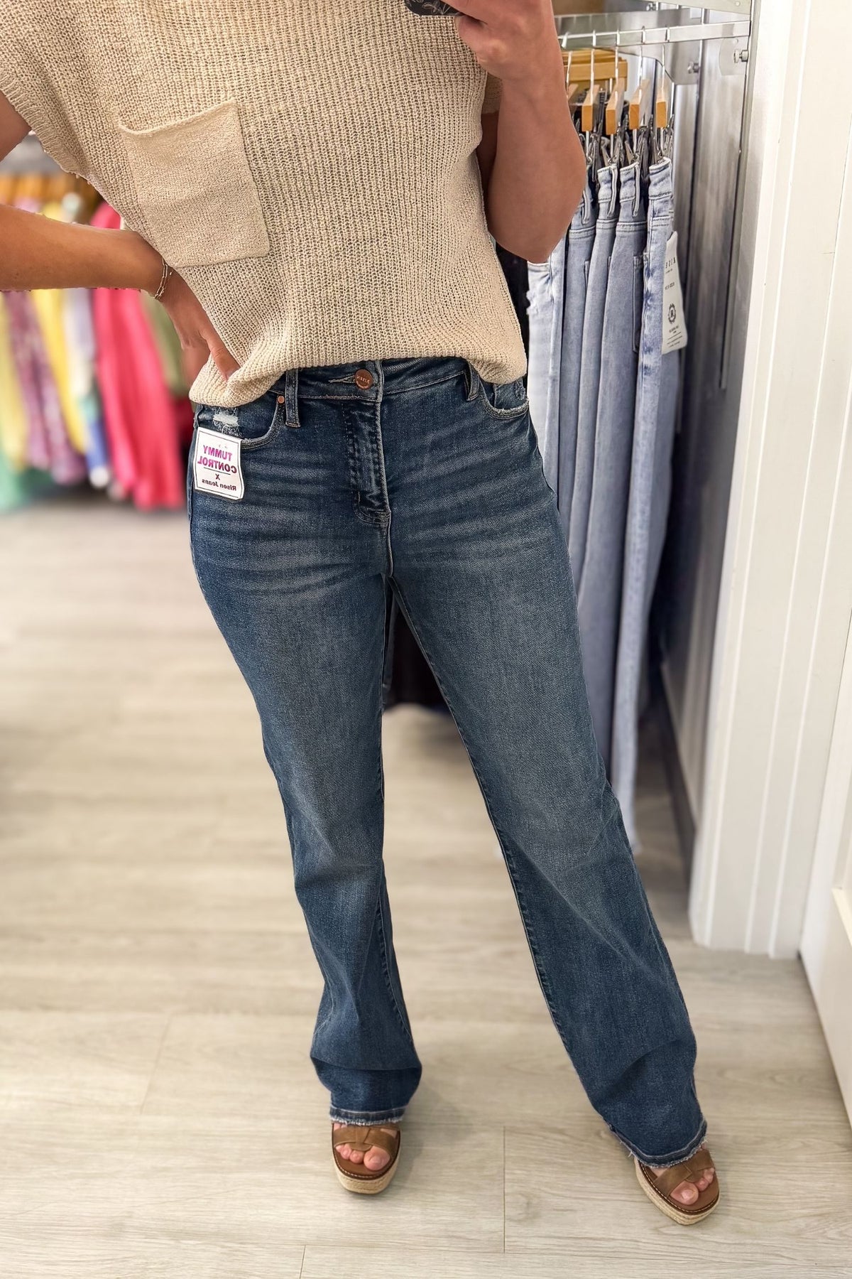 A person wearing a pair of dark-wash blue jeans with a straight leg, paired with a beige knit top and brown sandals, standing in a clothing store.