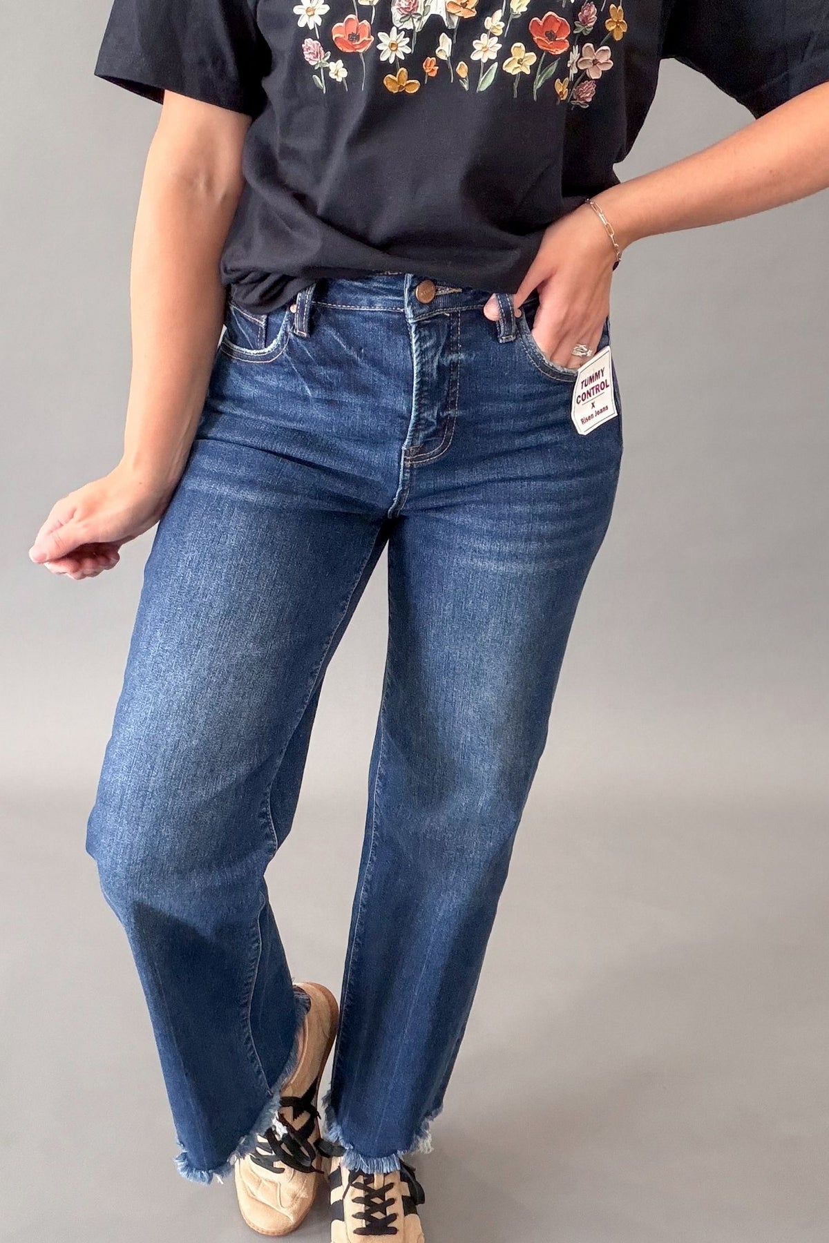 A person wearing dark-wash blue straight-leg jeans with a black graphic t-shirt and beige sneakers, standing against a plain gray studio background.