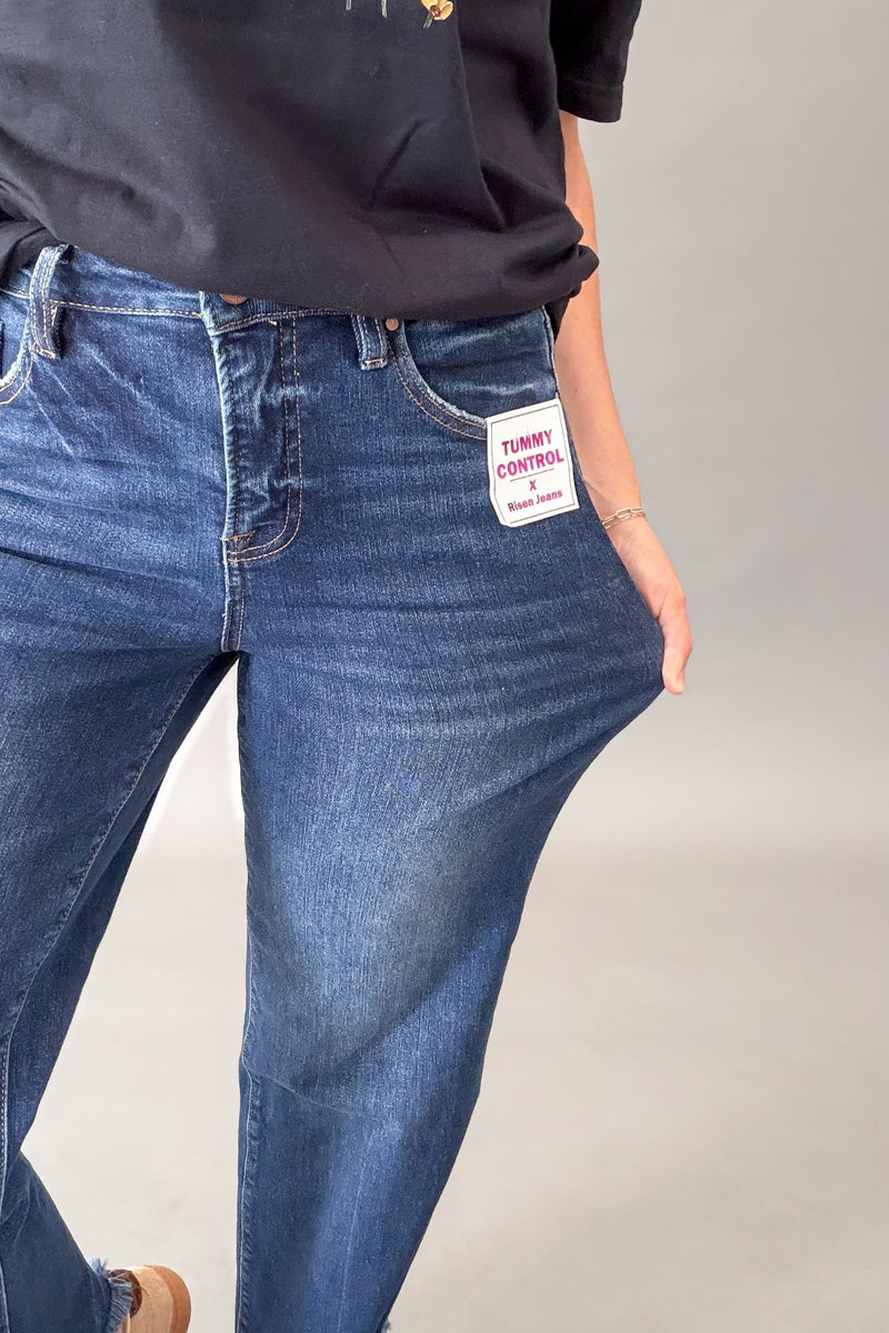 A close-up of a person wearing blue denim jeans with a stretchy side panel, pulling the fabric to demonstrate elasticity. A white patch on the pocket reads 'TUMMY CONTROL'.