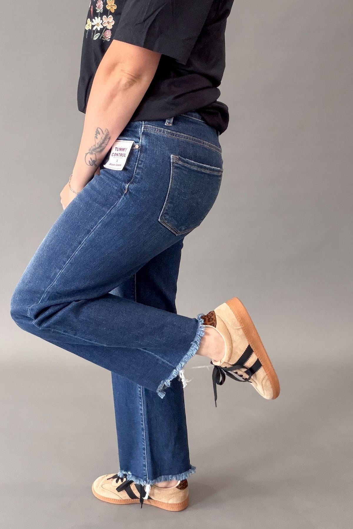 A person wearing blue denim jeans with a raw, frayed hem, styled with a black graphic t-shirt and beige-and-black sneakers shown from a side-rear angle against a solid gray studio background.