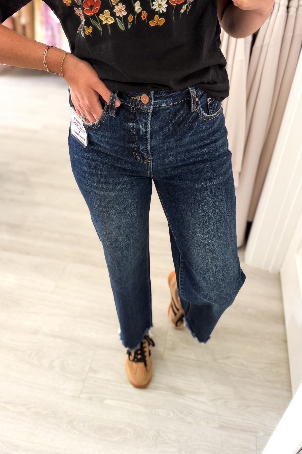 A person wearing dark blue straight-leg denim jeans with a faded wash, paired with a black graphic t-shirt and tan lace-up shoes, standing on a light wood floor.