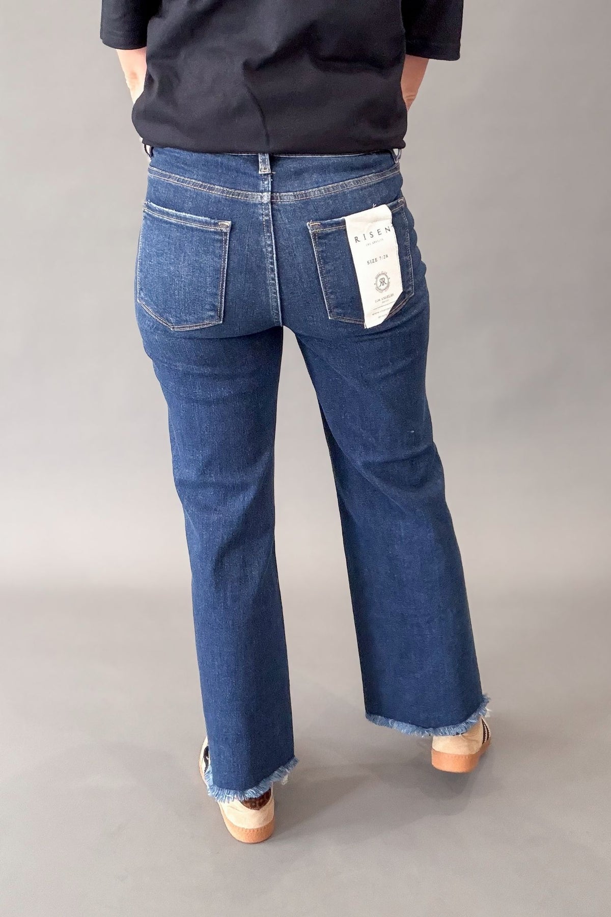 Rear view of a person wearing dark blue denim jeans with a raw frayed hem, paired with a black top and beige shoes, against a neutral gray studio background.