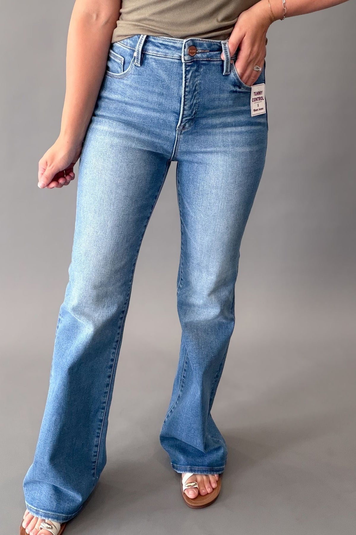 A person wearing medium blue high-rise Risen flare jeans with a faded wash, paired with a olive top and white slide sandals, shown against a neutral gray studio background.