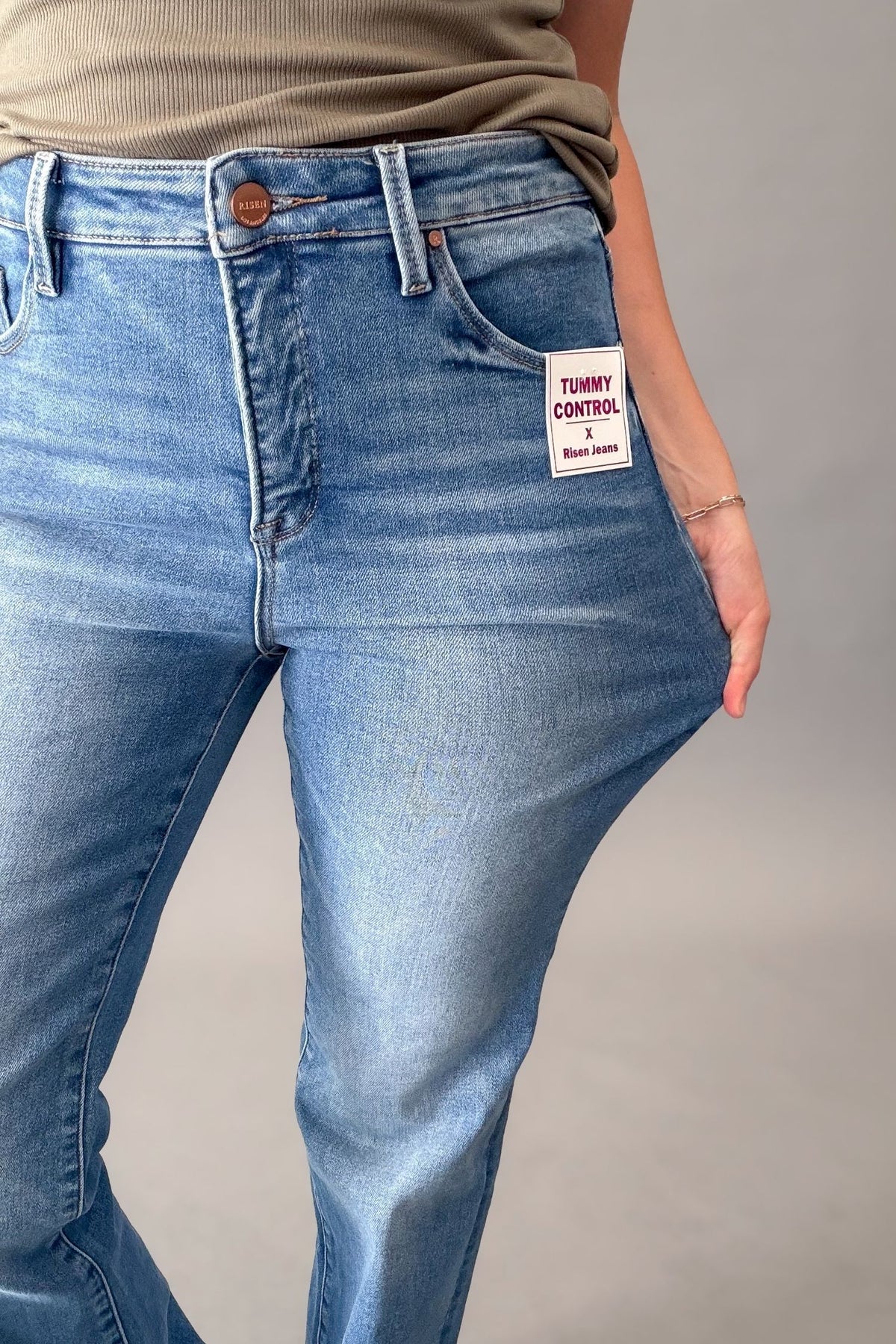 Close-up of a person wearing medium blue risen denim jeans with a visible front patch reading 'TUMMY CONTROL'. The photo is cropped from the waist down against a plain light gray studio background.