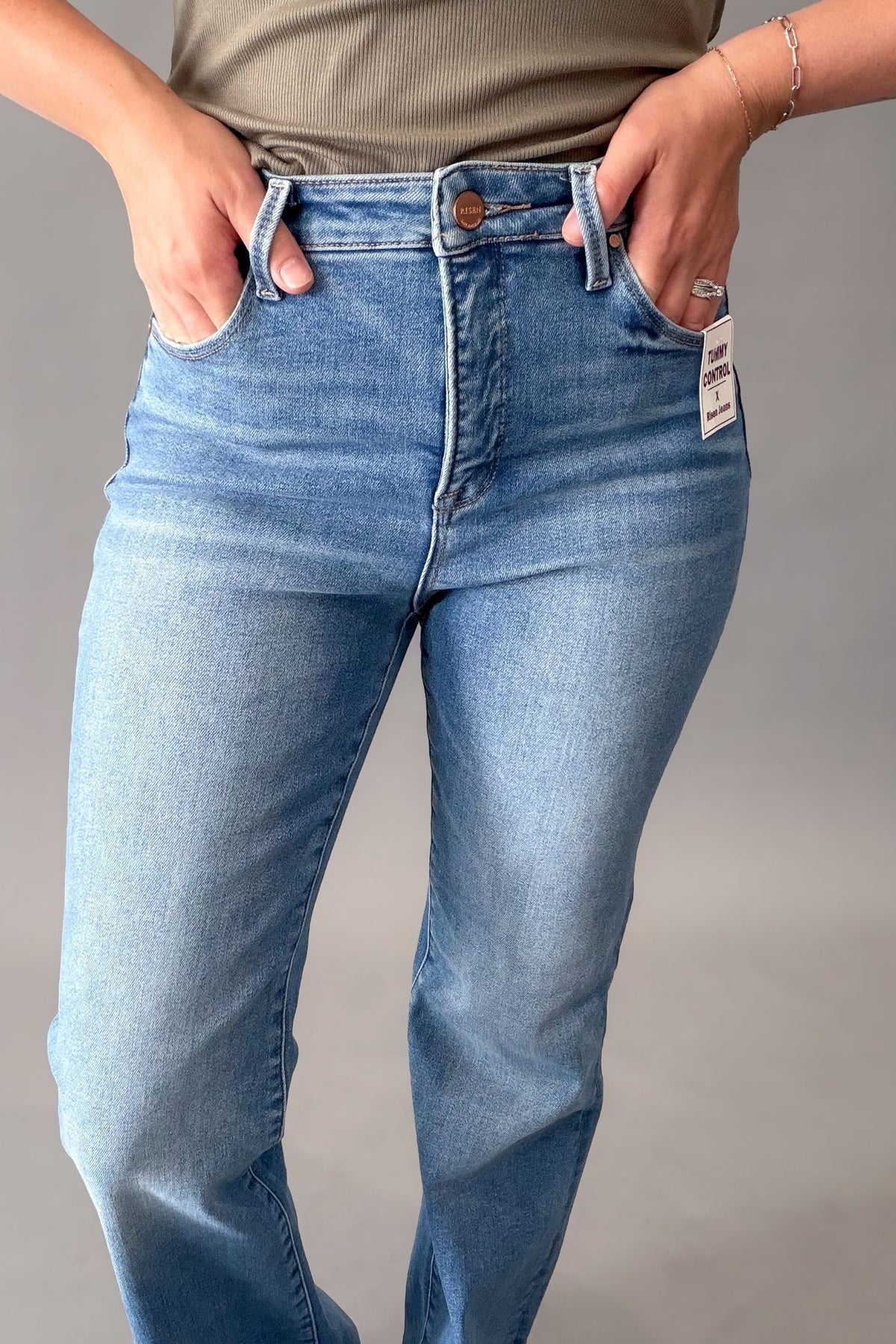A person wearing medium blue denim jeans with a visible white size tag on the pocket, standing against a plain gray studio background.