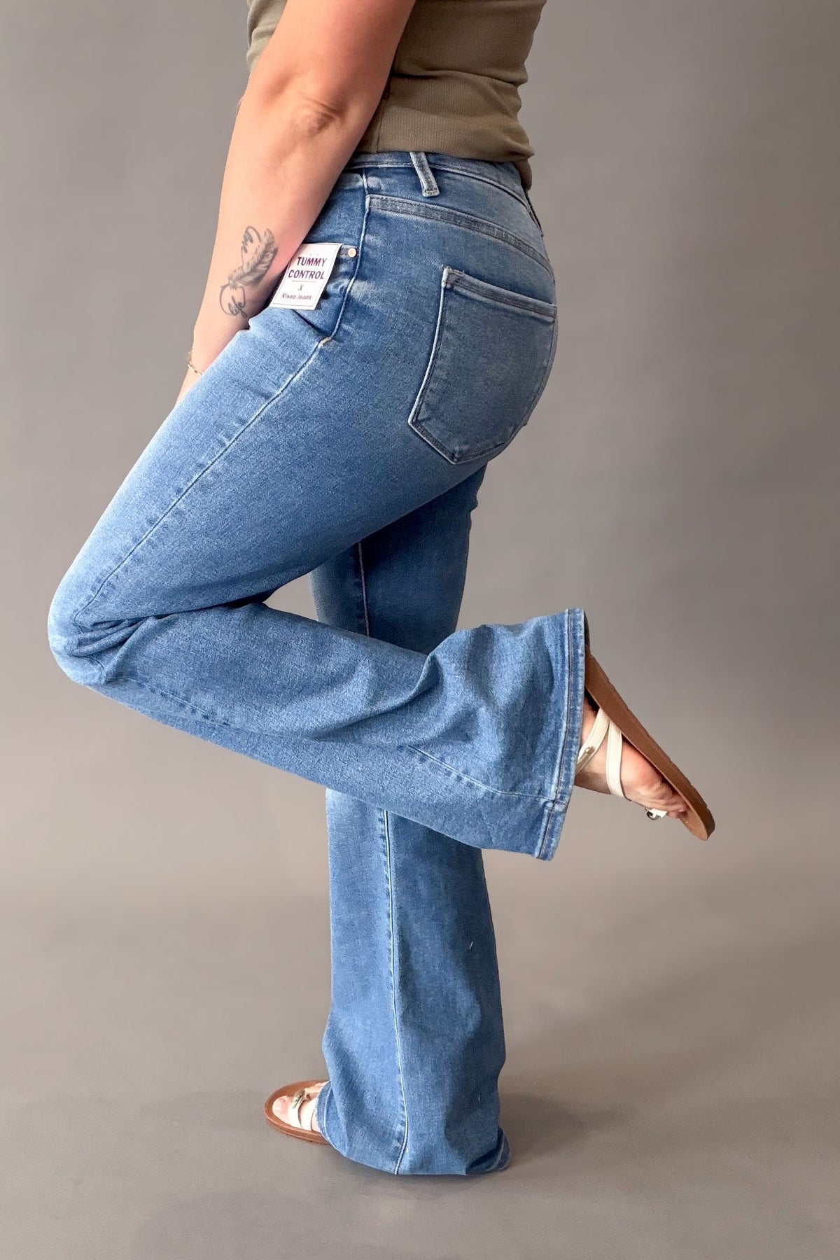 A person wearing medium blue flared jeans, standing in a studio against a plain gray background. The photo shows the back and side of the jeans, with one leg bent upward.