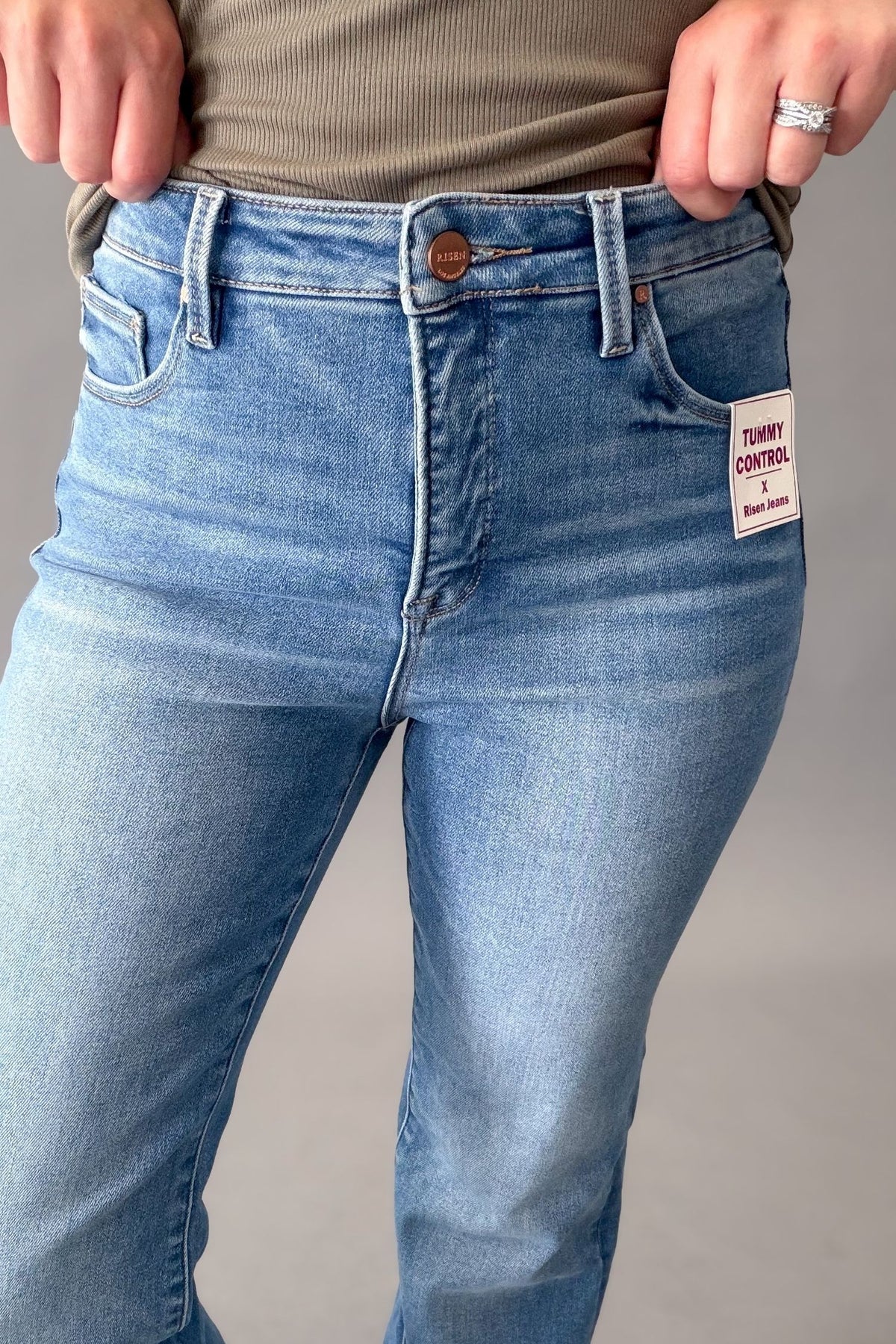 Close-up of a person wearing medium blue Tummy control risen jeans, showing the front waistband, button, belt loops, front pockets, and a side brand patch. The person is also wearing a olive ribbed top against a plain light gray studio background.