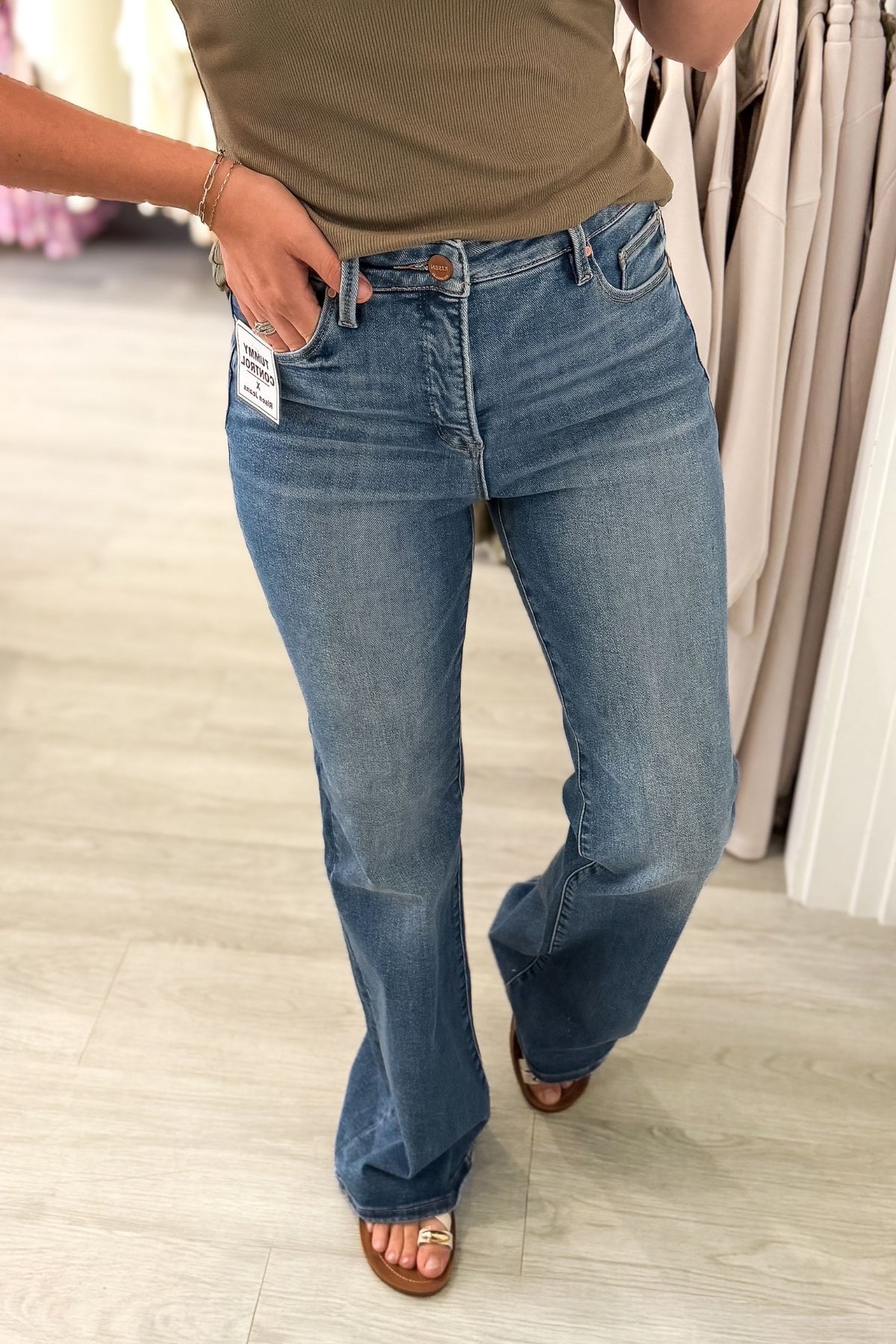A person wearing medium-wash blue flare jeans with a white brand tag on the pocket, paired with a olive top and brown sandals in a retail store setting.