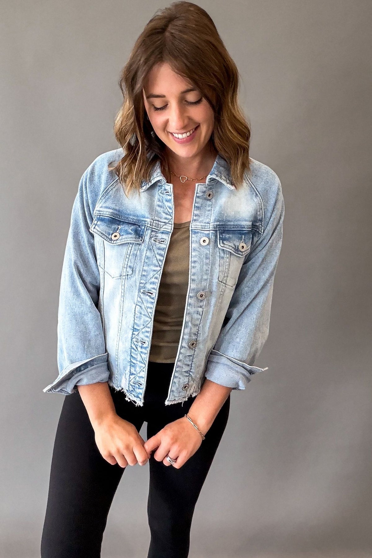 A woman wearing a light-wash blue denim jacket over a brown top and black pants, standing against a plain gray studio background.